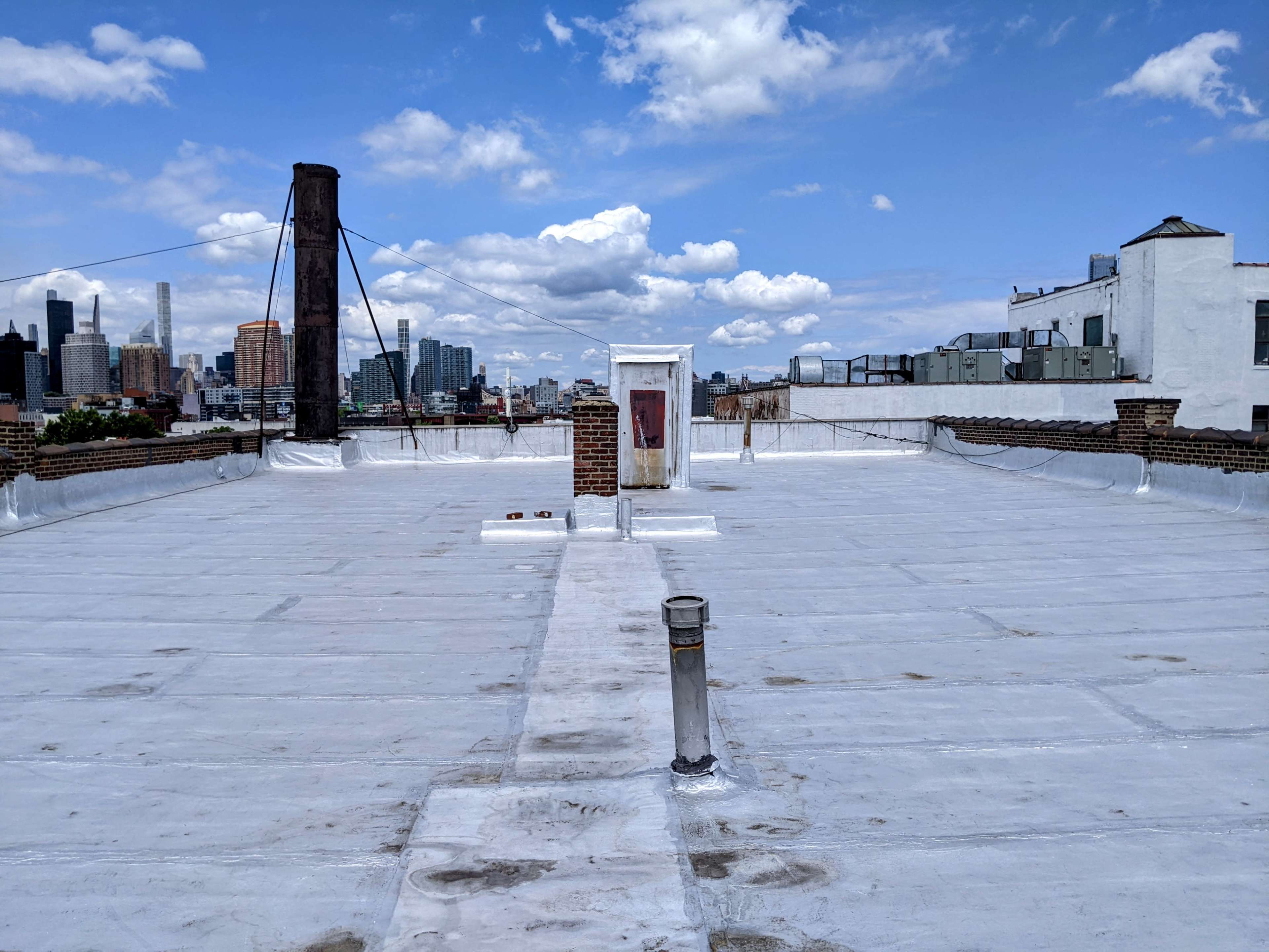 A flat rooftop with a white surface and a central access door overlooks a city skyline under a partly cloudy sky.