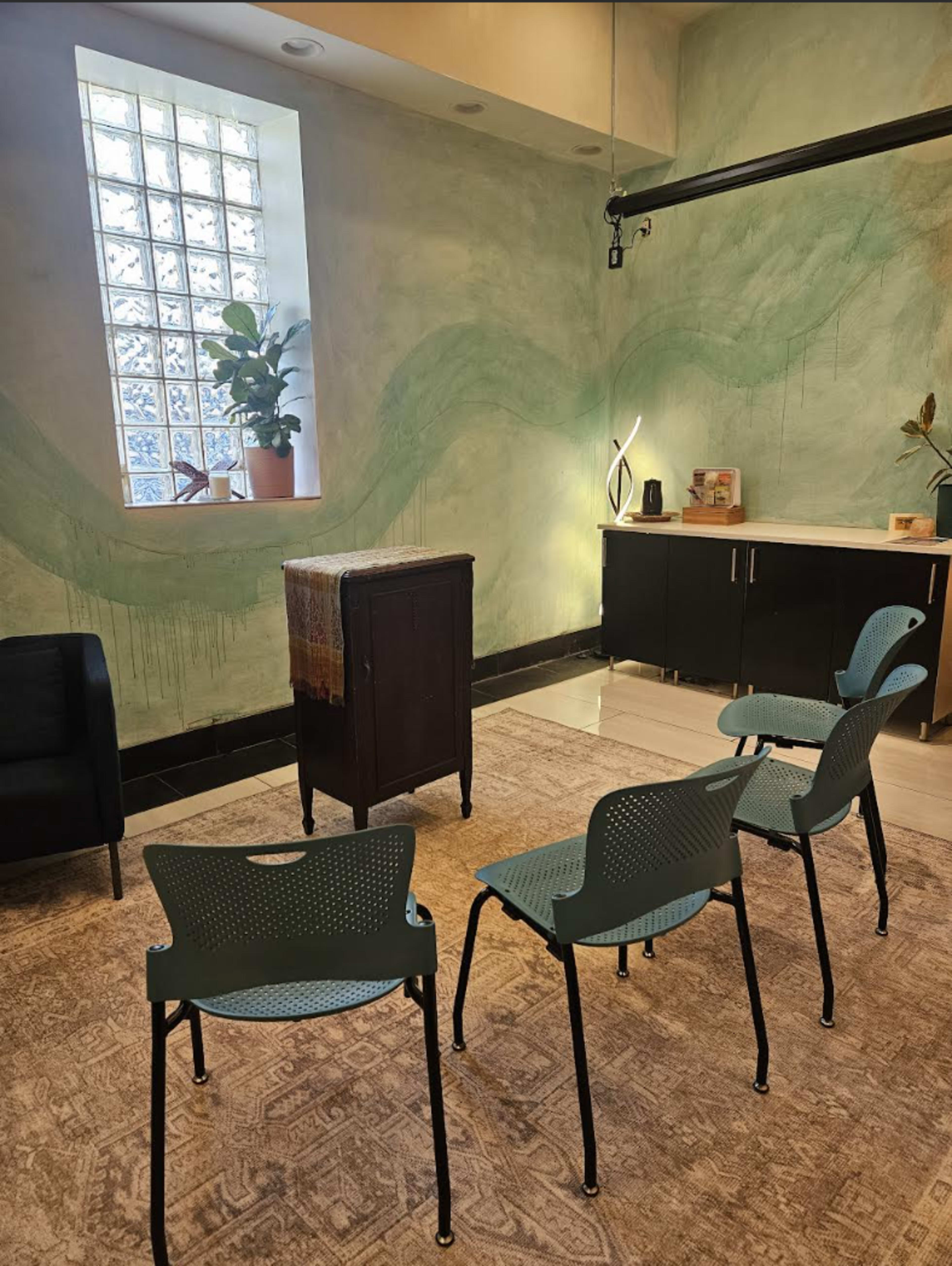 The image shows a modern waiting area with four blue chairs arranged in front of a black cabinet, set against a painted green wall and illuminated by natural light from a nearby window.