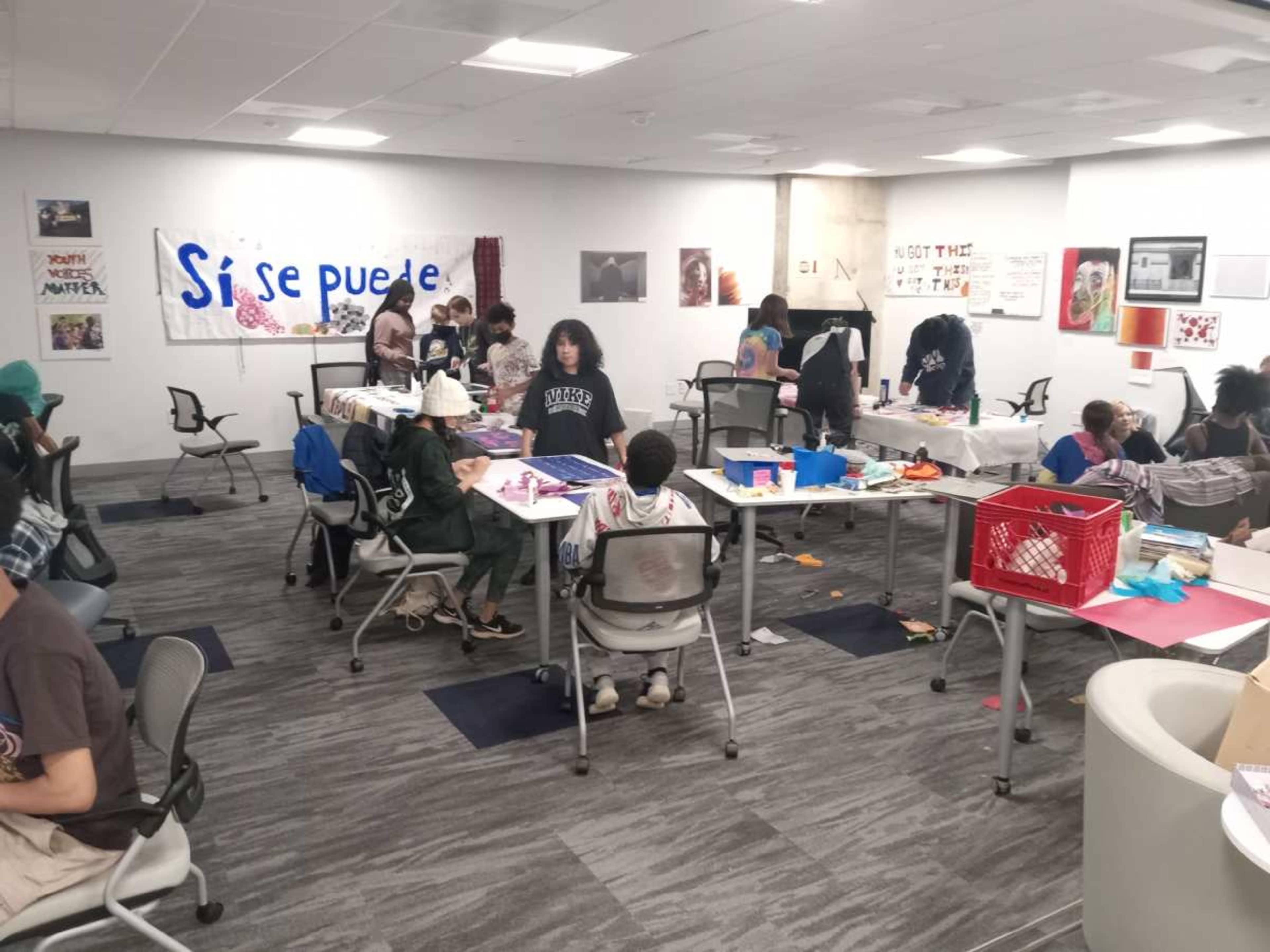 A group of children and adults engage in an art-making activity in a brightly lit community space with tables and various materials scattered around.