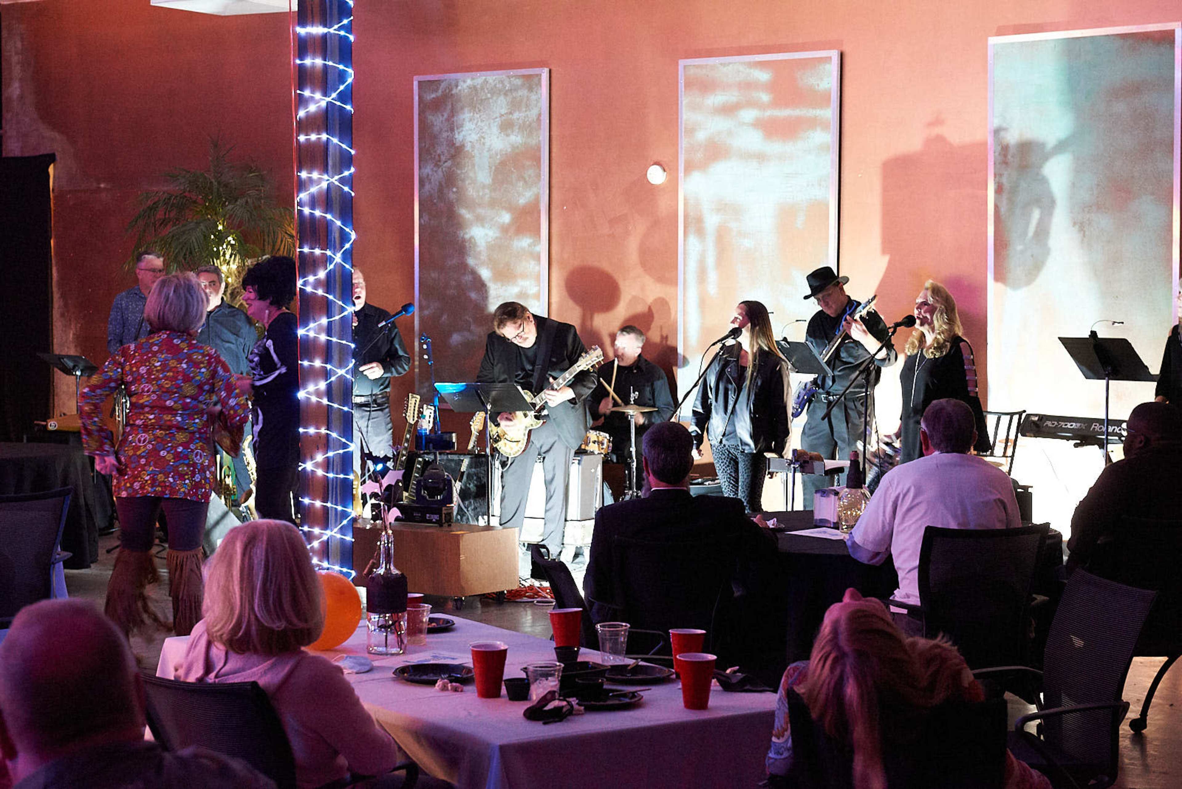 A live band performs on stage in a decorated venue with scattered tables and attendees enjoying the event.