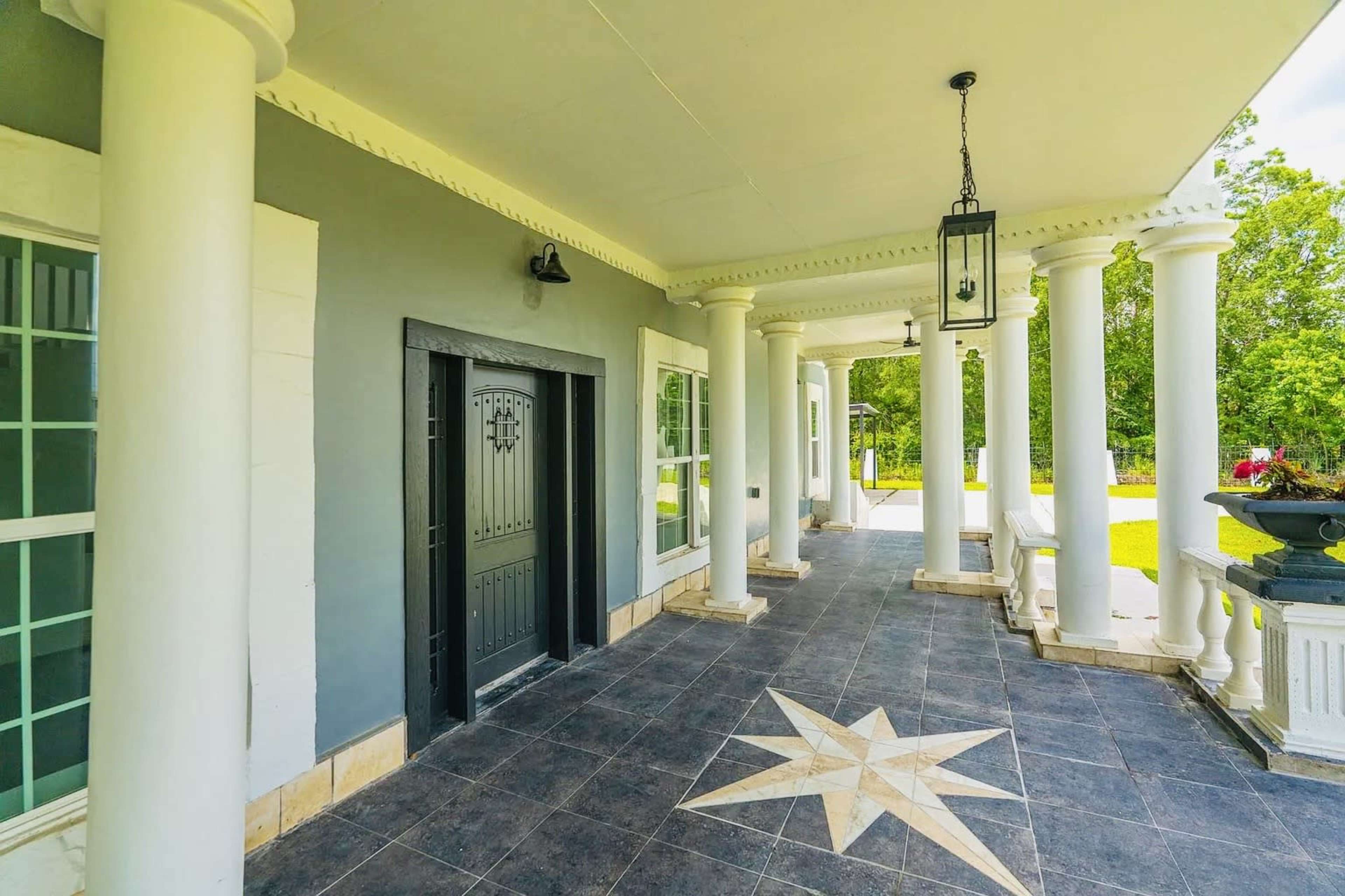 The image shows a covered walkway with large columns, a decorative star design on the floor, and a black door leading to a building.