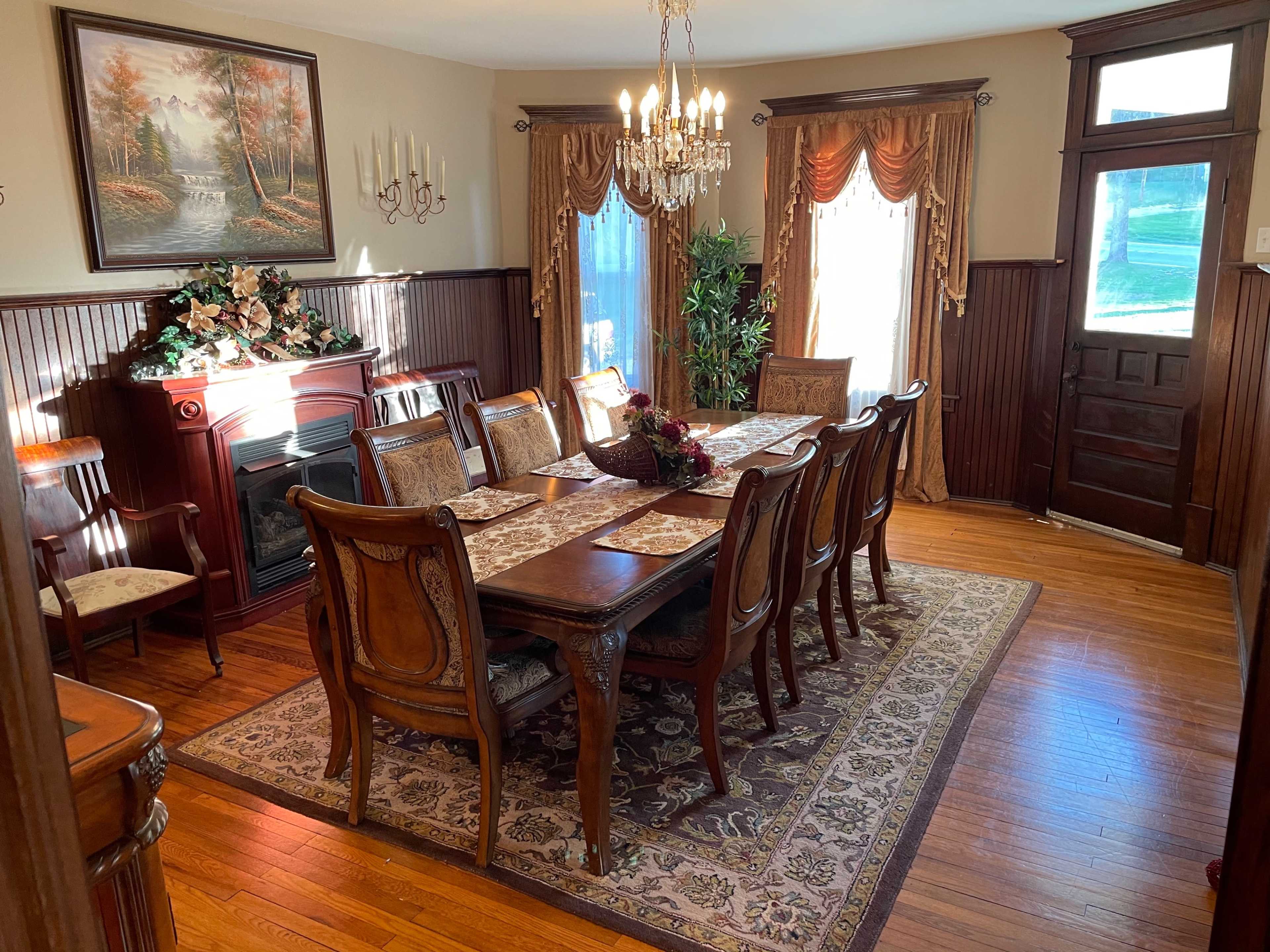 A dining room with a large wooden table set for a meal, surrounded by upholstered chairs, and featuring decorative elements such as a chandelier, a fireplace, and a large painting on the wall.