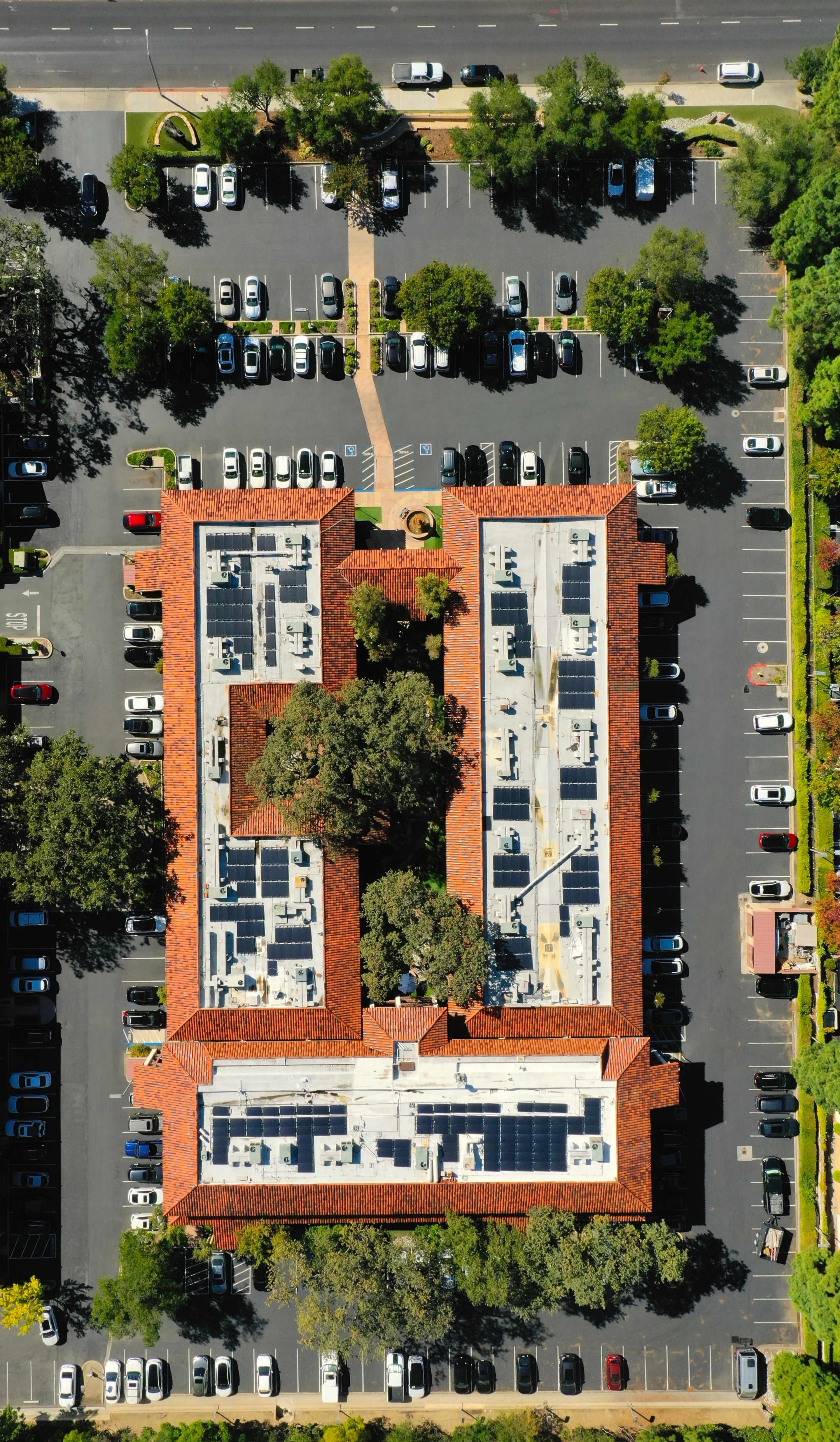 The image shows an aerial view of a building with a red-tiled roof surrounded by a parking lot filled with vehicles and lined with trees.