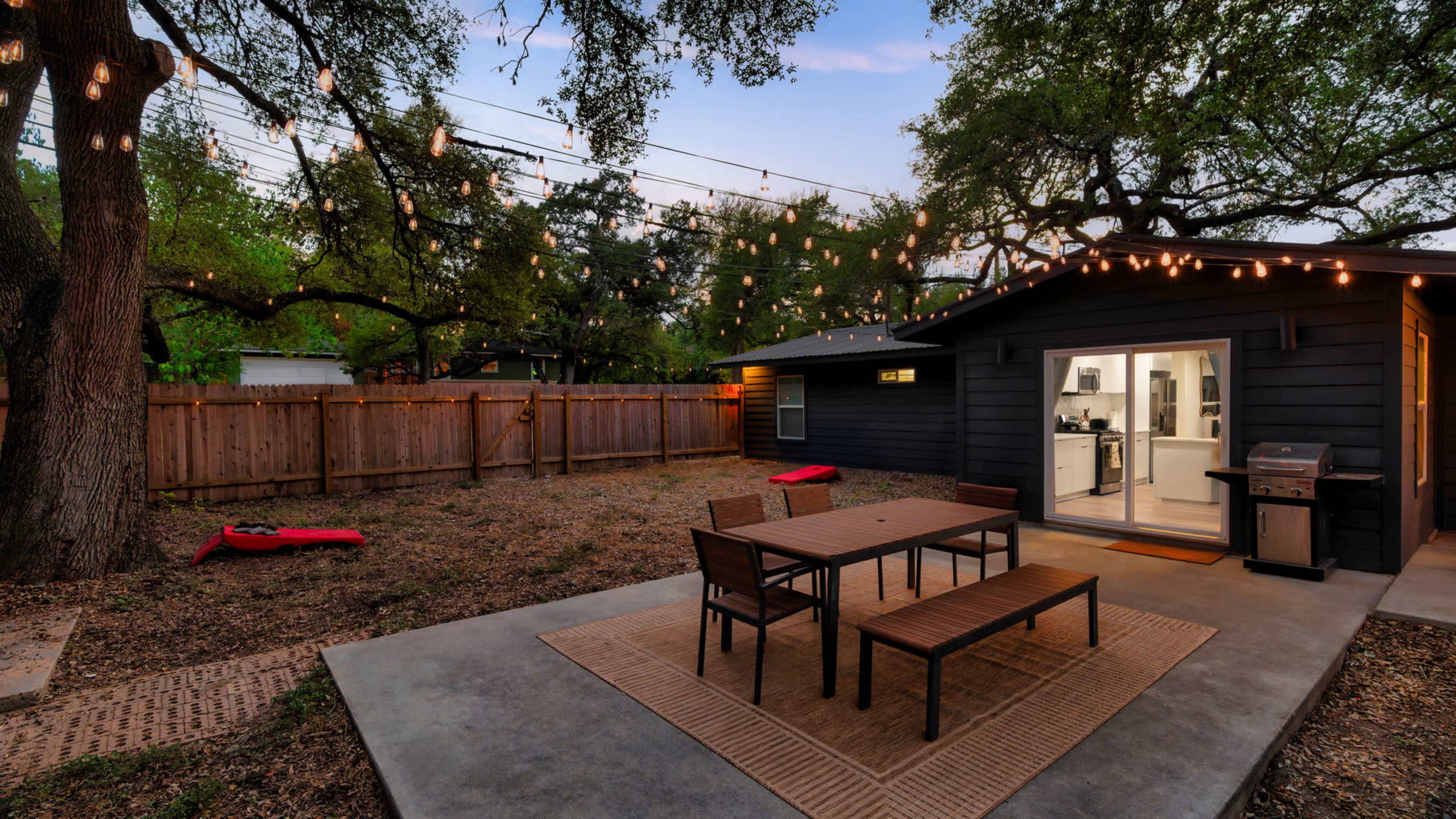 A cozy outdoor patio features a table and chairs surrounded by string lights, leading to a modern kitchen visible through a glass door.