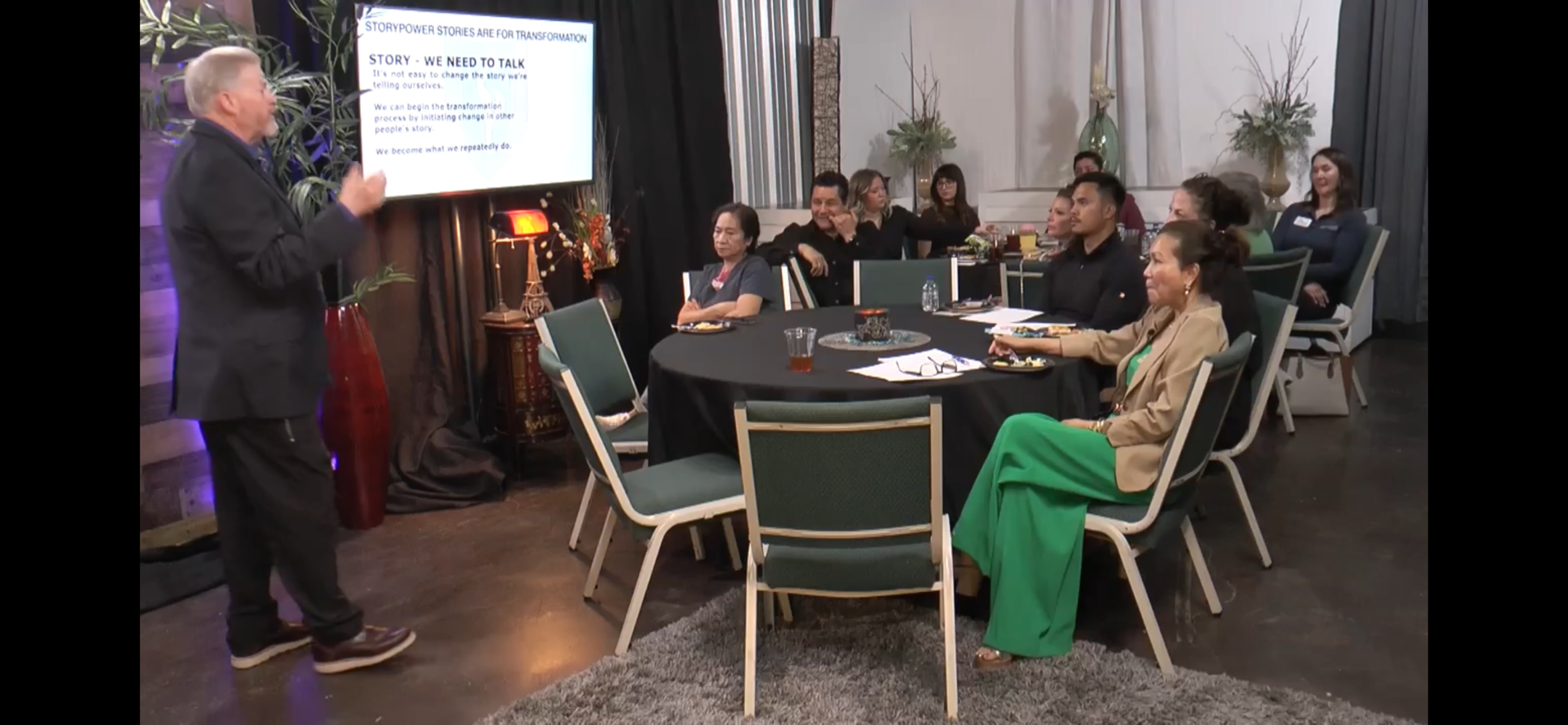 A speaker presents to an audience seated at round tables in a well-lit room with plants and decorative elements.