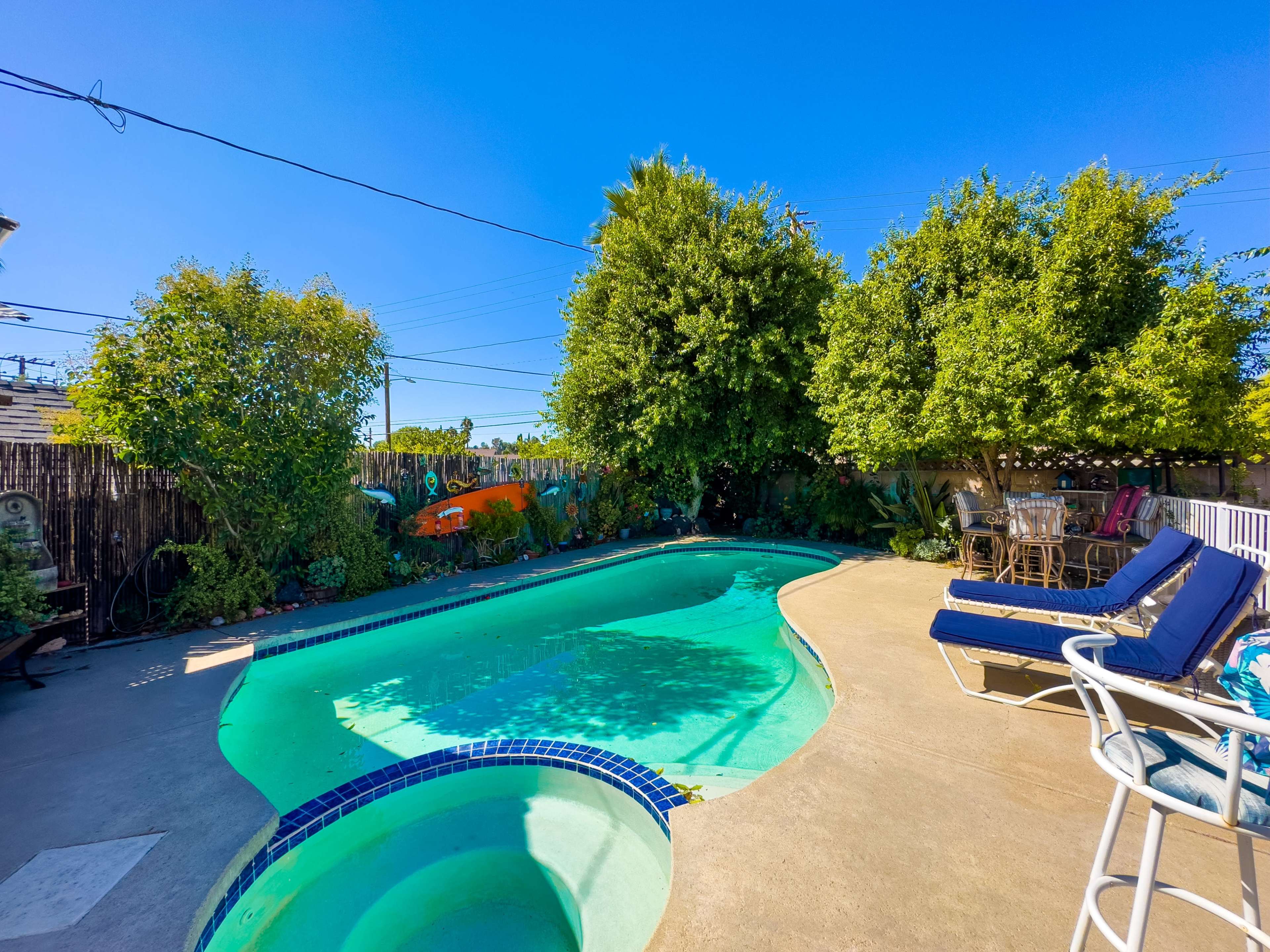 The image shows a residential backyard with a swimming pool surrounded by greenery and a seating area.