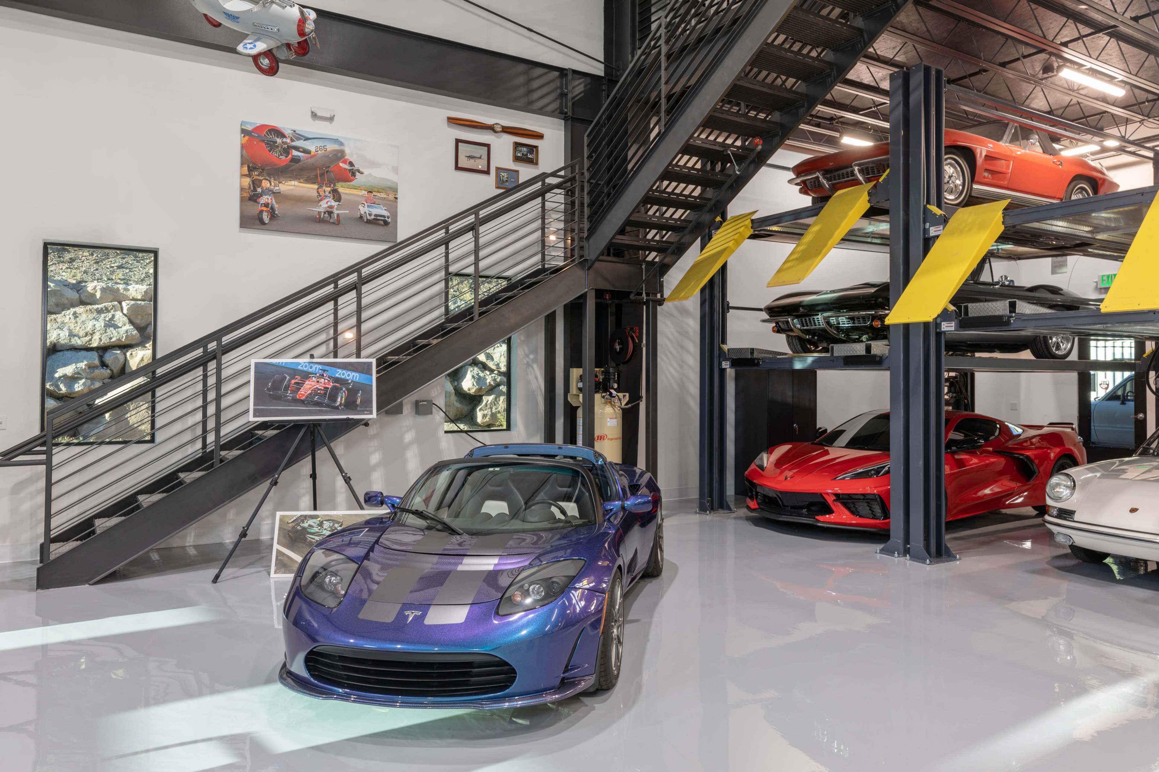 A modern garage features a blue sports car and a red sports car displayed on lifts, with art pieces and collectibles arranged on the walls.