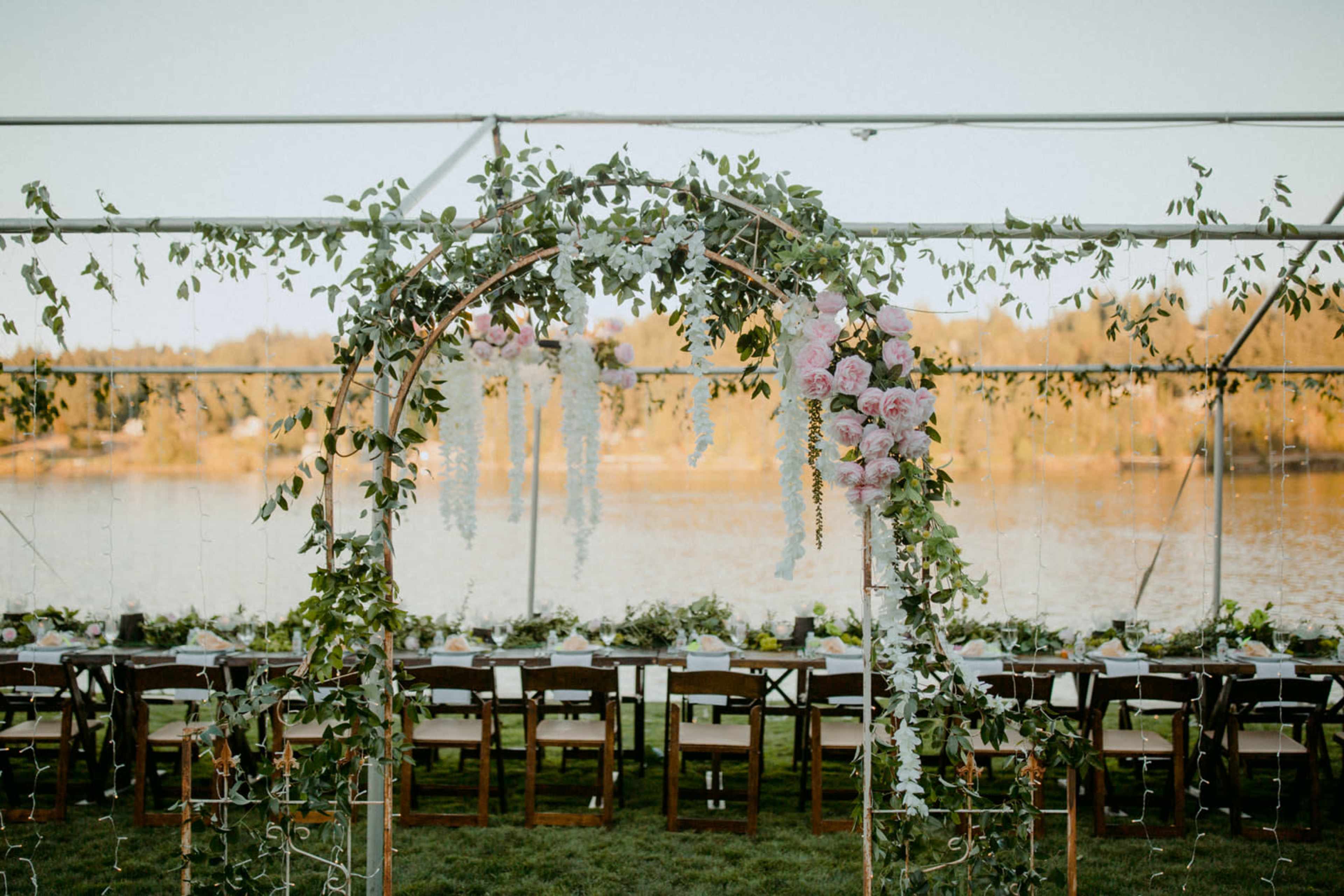 A wooden arch adorned with flowers and greenery stands in front of a long table set for a banquet beside a serene lake.