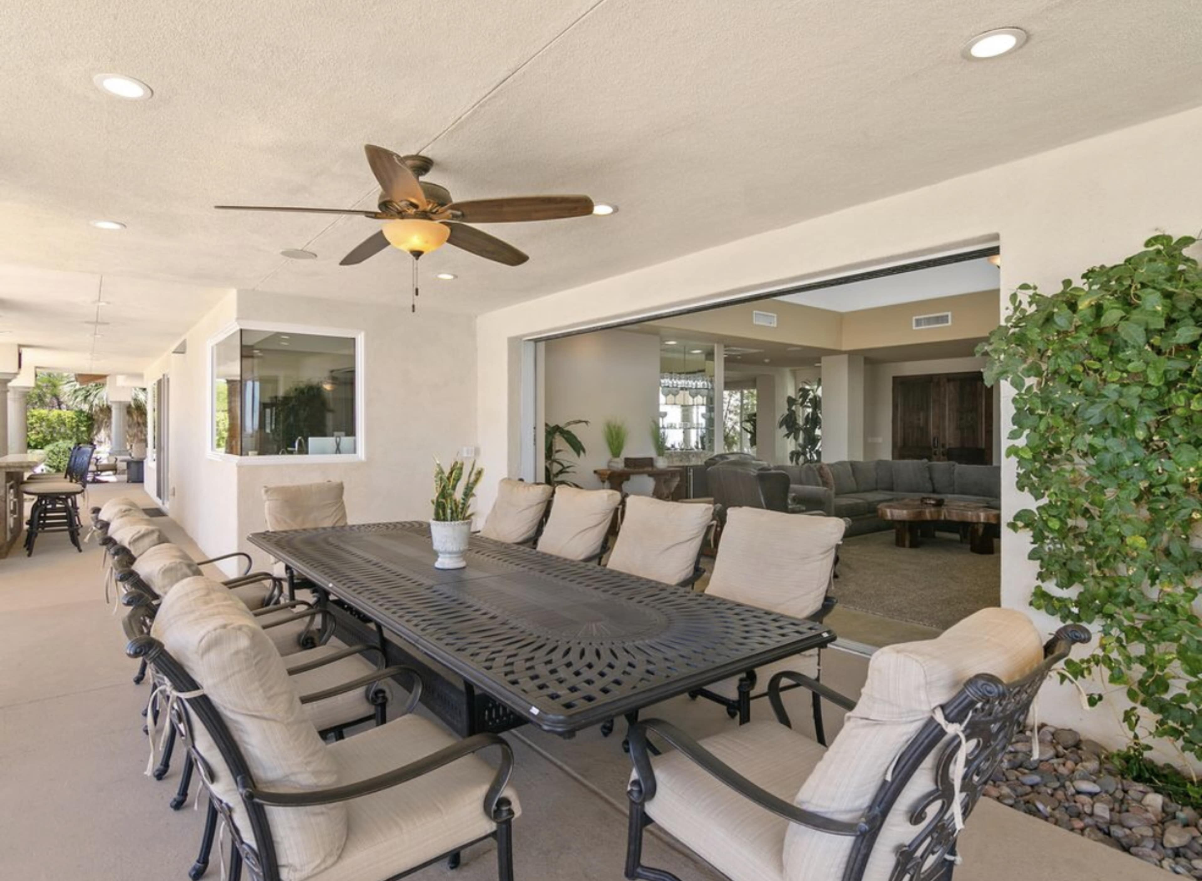 A covered patio area featuring a large dining table surrounded by chairs, with a ceiling fan above and a view of an interior living space through large glass windows.