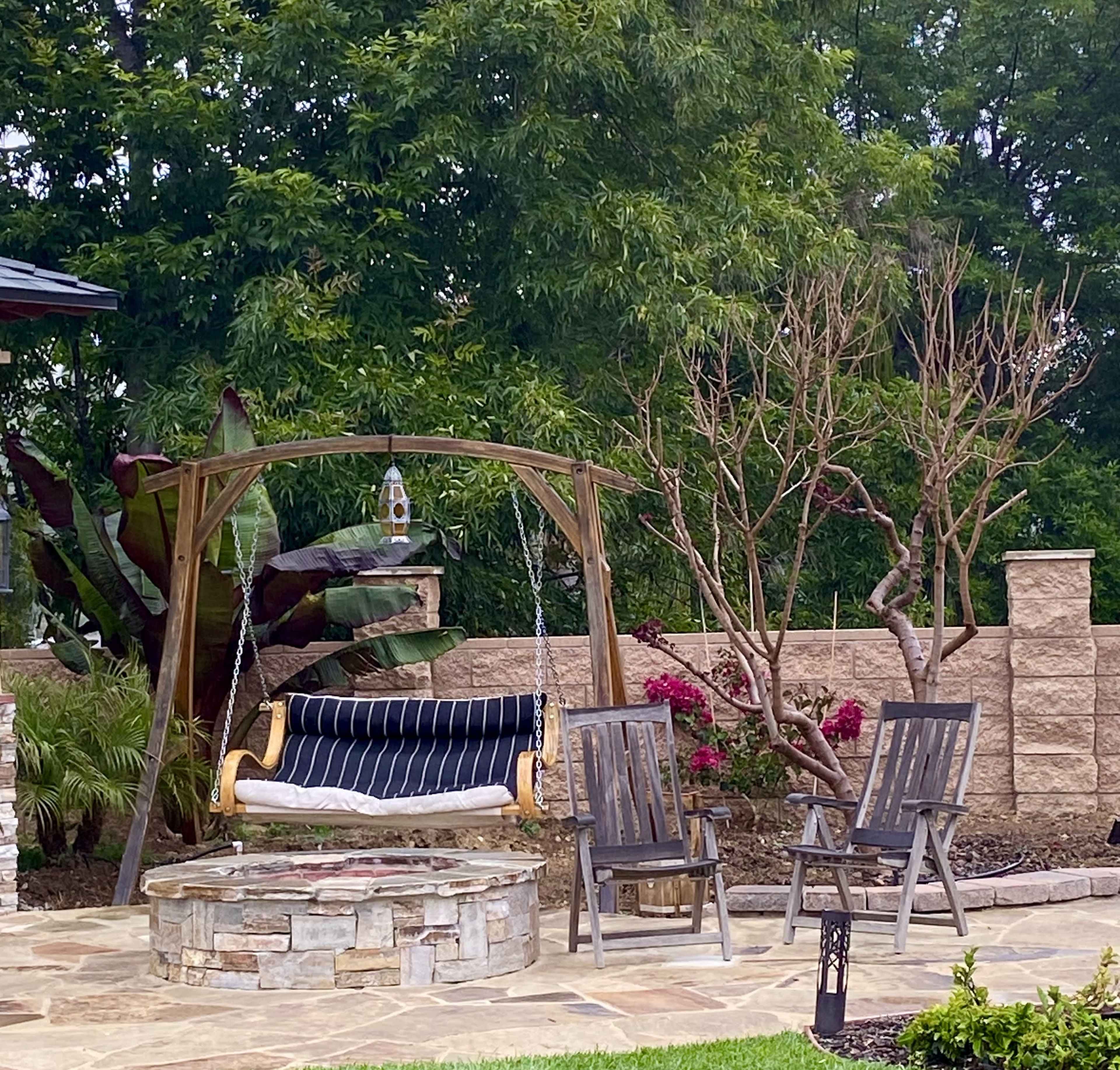 A stone fire pit surrounded by two wooden chairs and a swing on a wooden frame in a landscaped garden with green plants and a flowering bush.