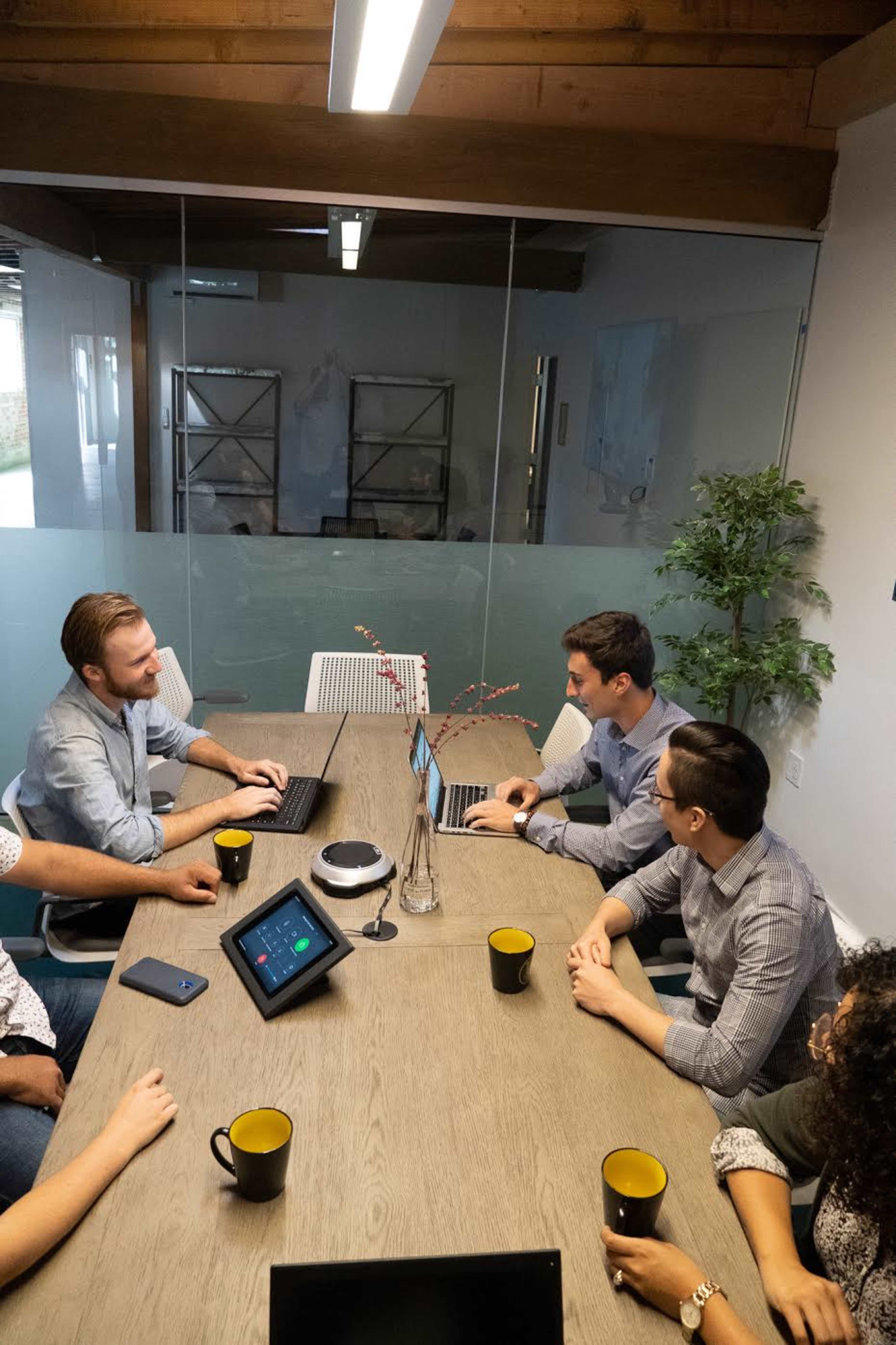 A group of six people are seated around a wooden table in a conference room, engaged in a discussion while using laptops and tablets.