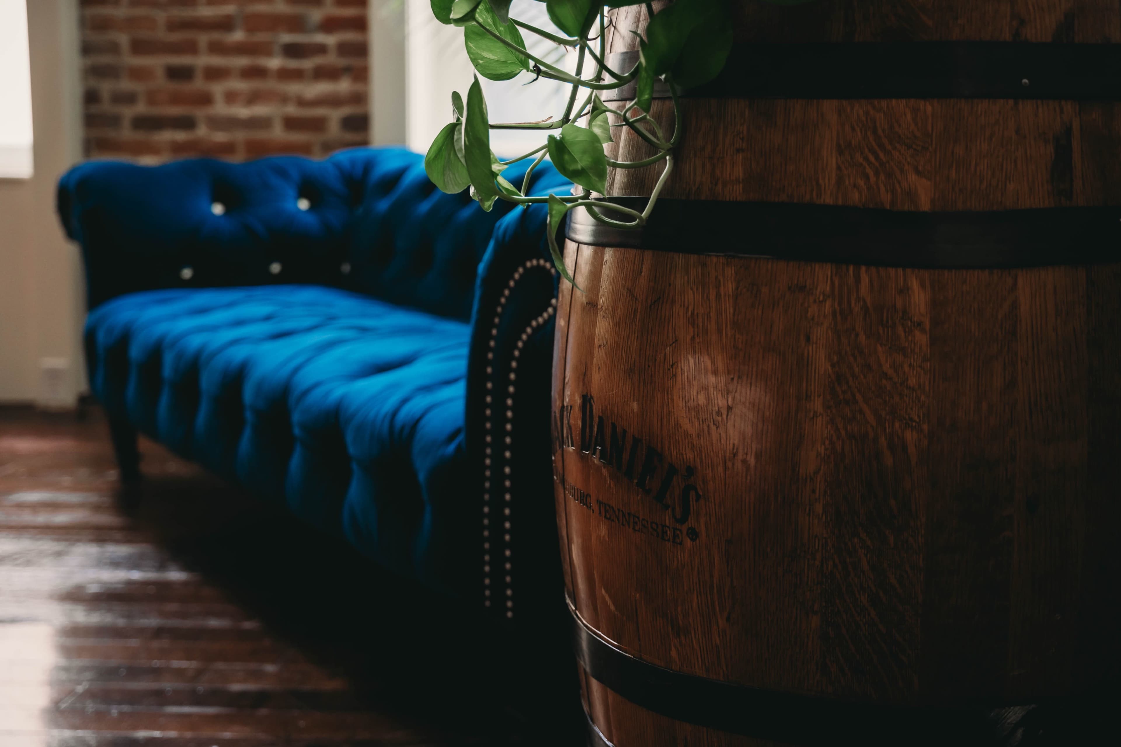 A blue tufted sofa is positioned next to a wooden barrel with a plant emerging from it in a rustic interior.