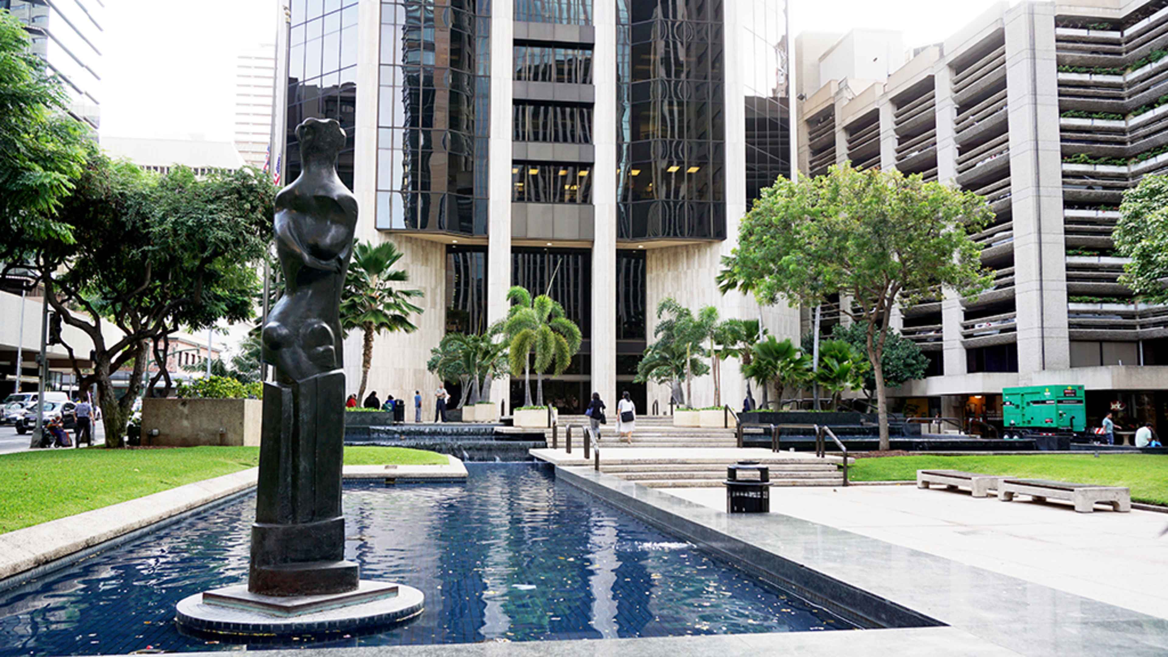 A tall statue is positioned beside a reflecting pool in a landscaped area in front of a modern glass building and a parking structure.