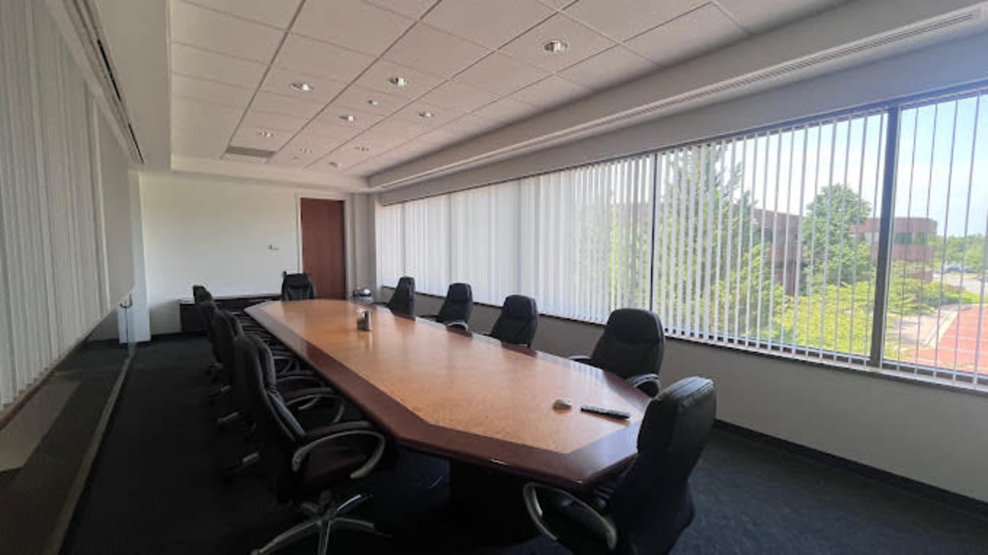 A conference room features a long table surrounded by black office chairs, with large windows allowing natural light to fill the space.