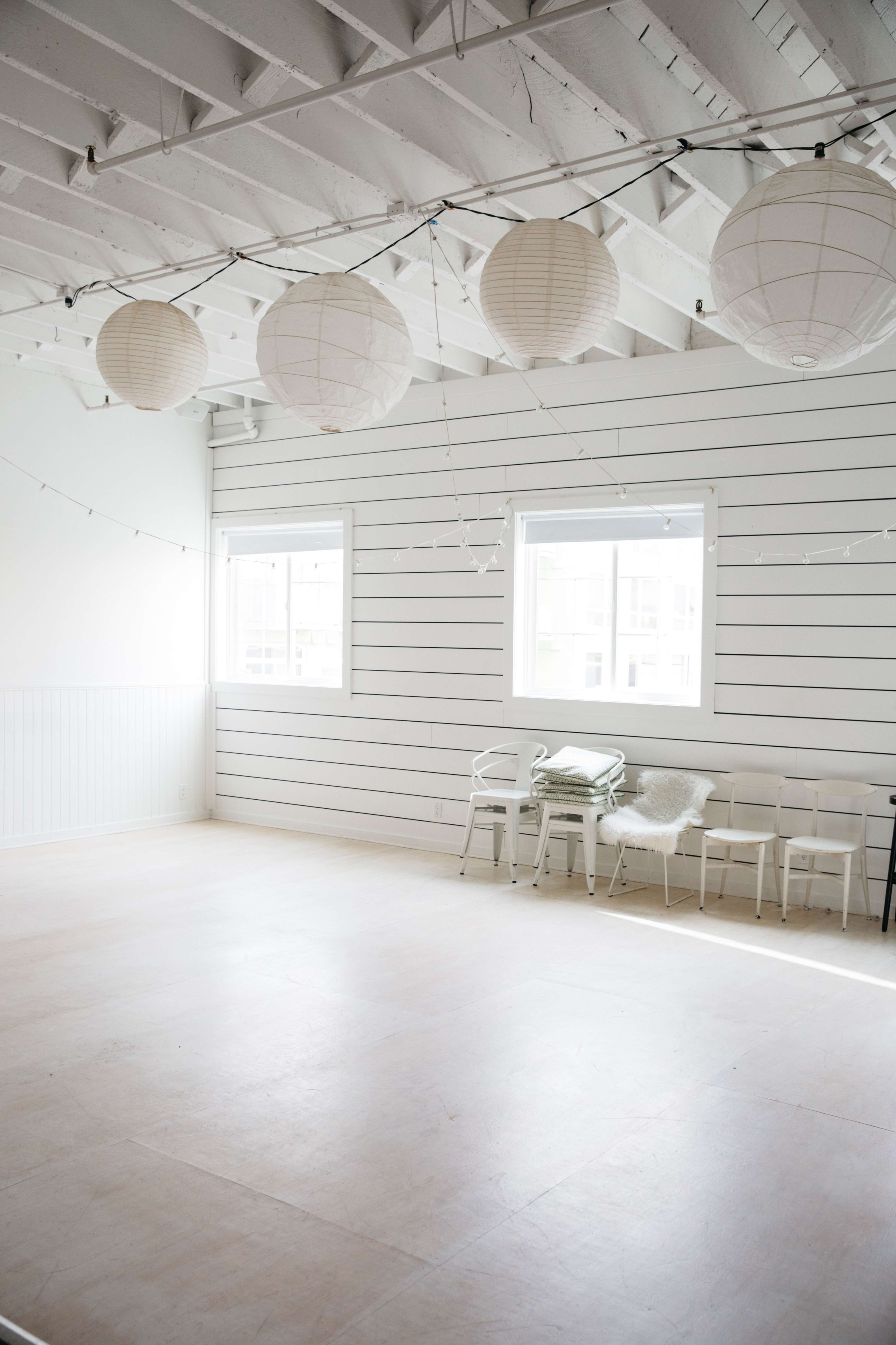 A bright, empty room features white walls with wooden paneling, overhead paper lanterns, and several chairs lined against the wall.
