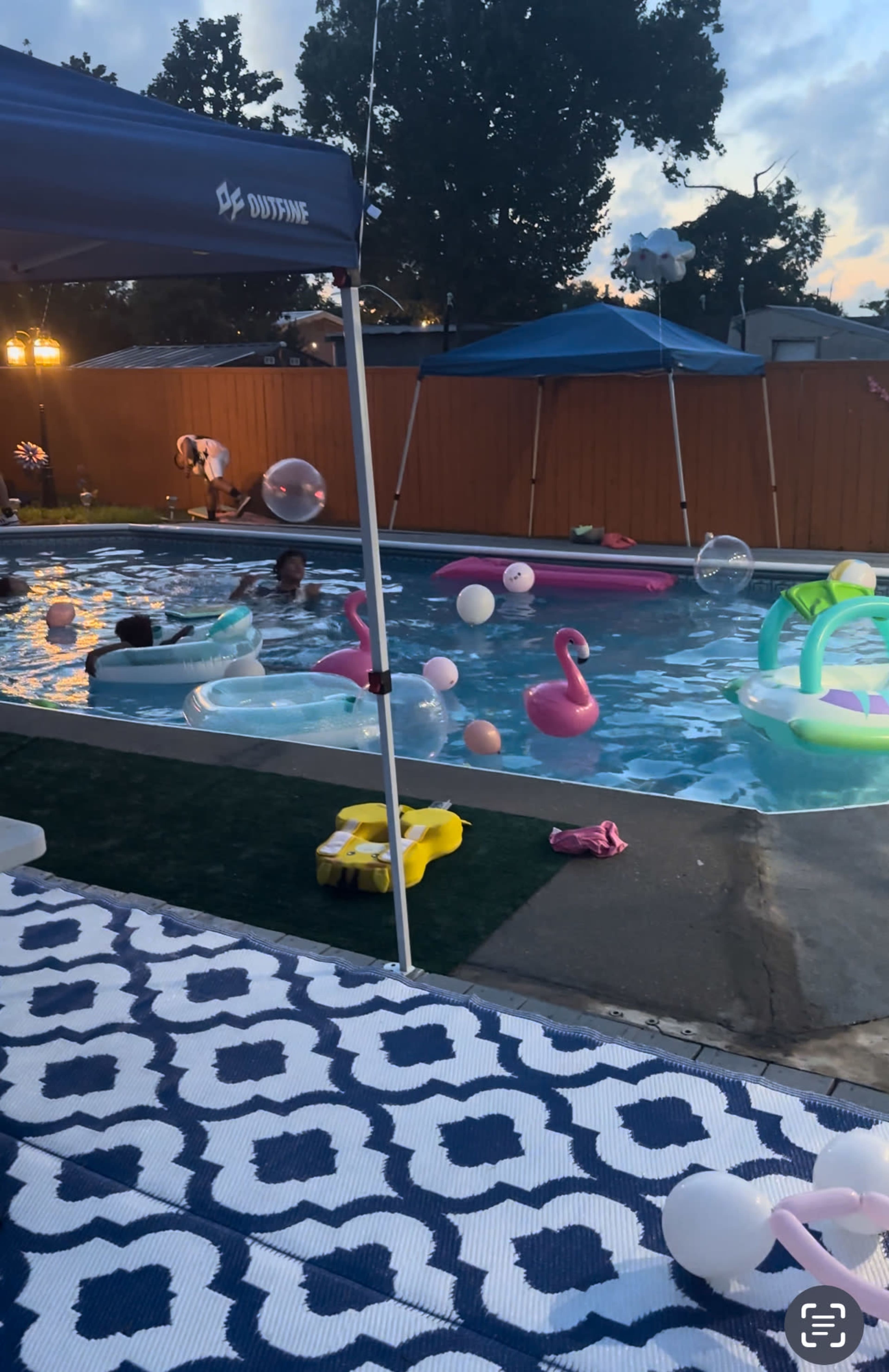 The image shows a backyard pool filled with inflatable floats and people enjoying the water, under a tent with decorative balloons nearby.