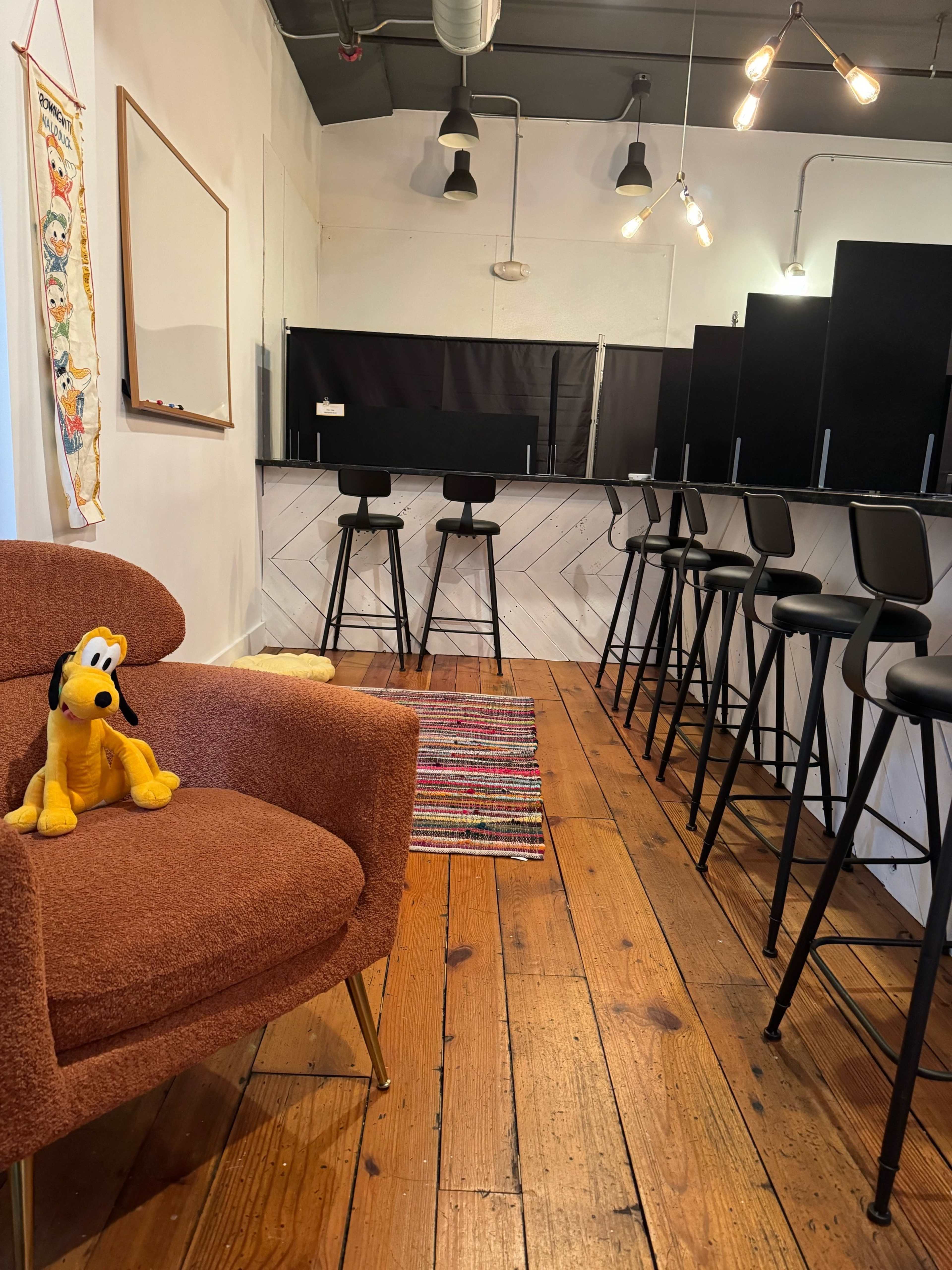 A spacious room with wooden floors, featuring a cozy brown chair and a bar area with black stools along a white wall.