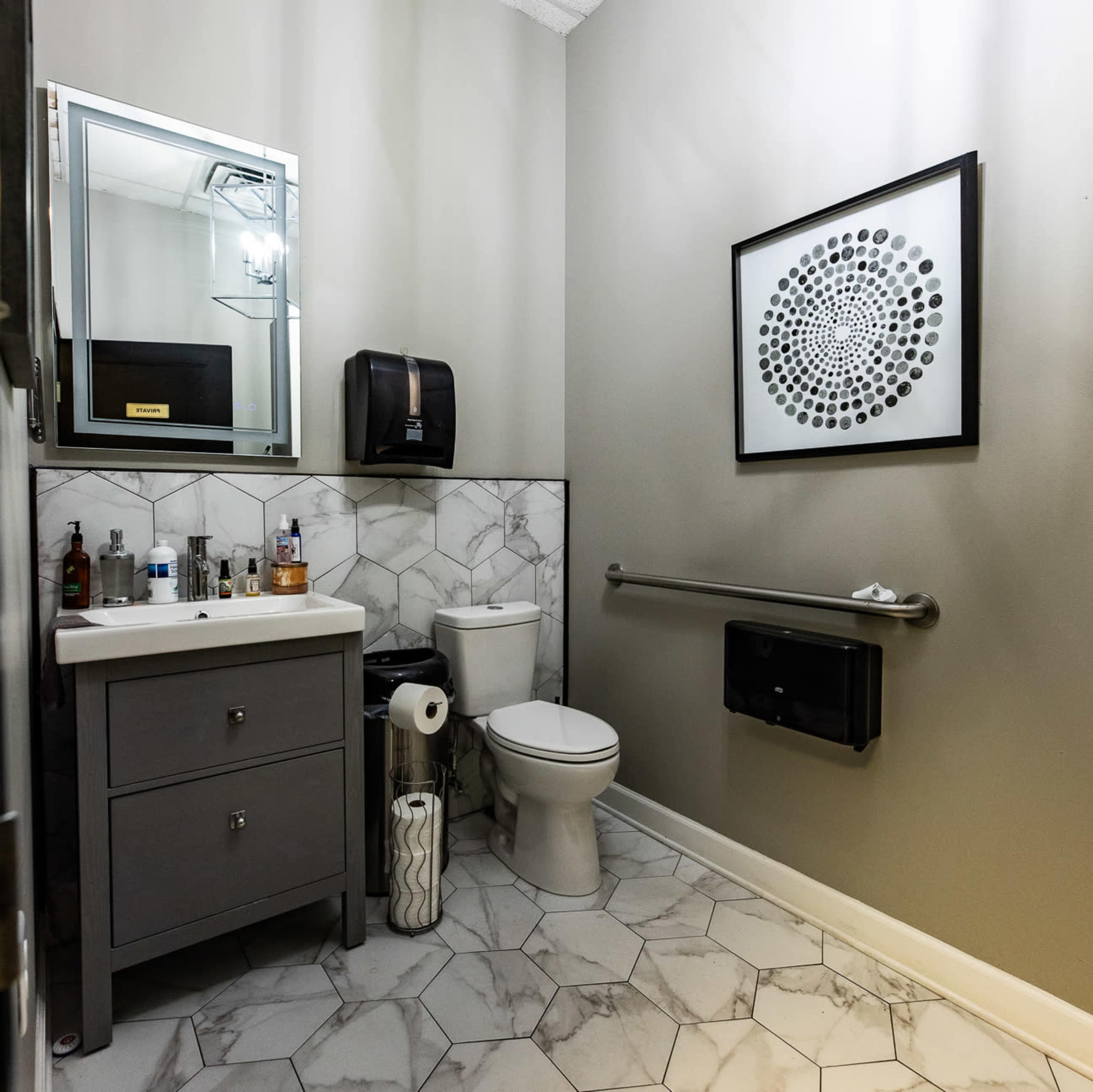 The image shows a small bathroom featuring a white toilet, a gray vanity with a sink, and tiled flooring with a hexagonal pattern.