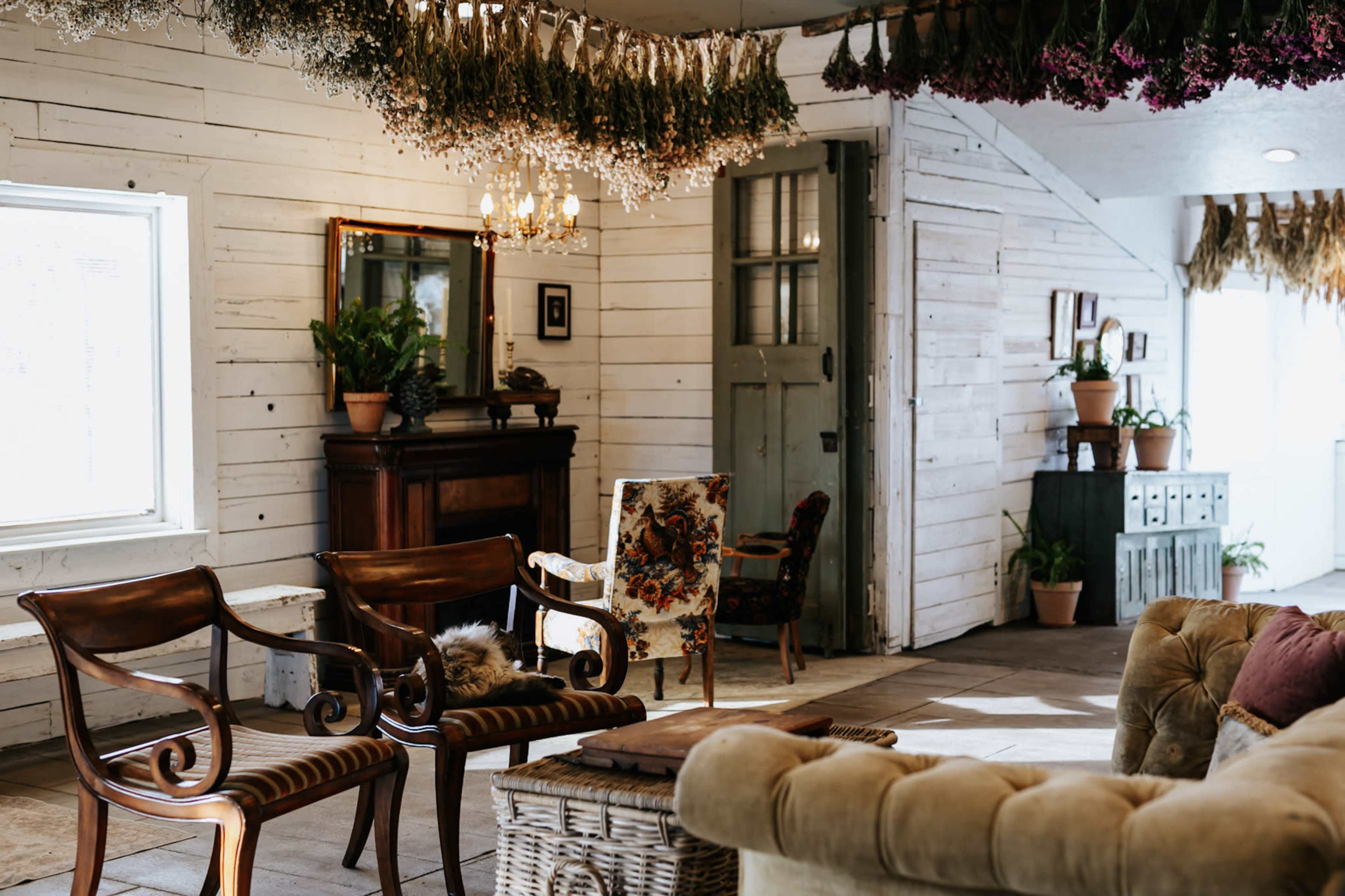 The image shows a rustic living space featuring vintage wooden furniture, decorative plants, and a variety of hanging dried flowers.