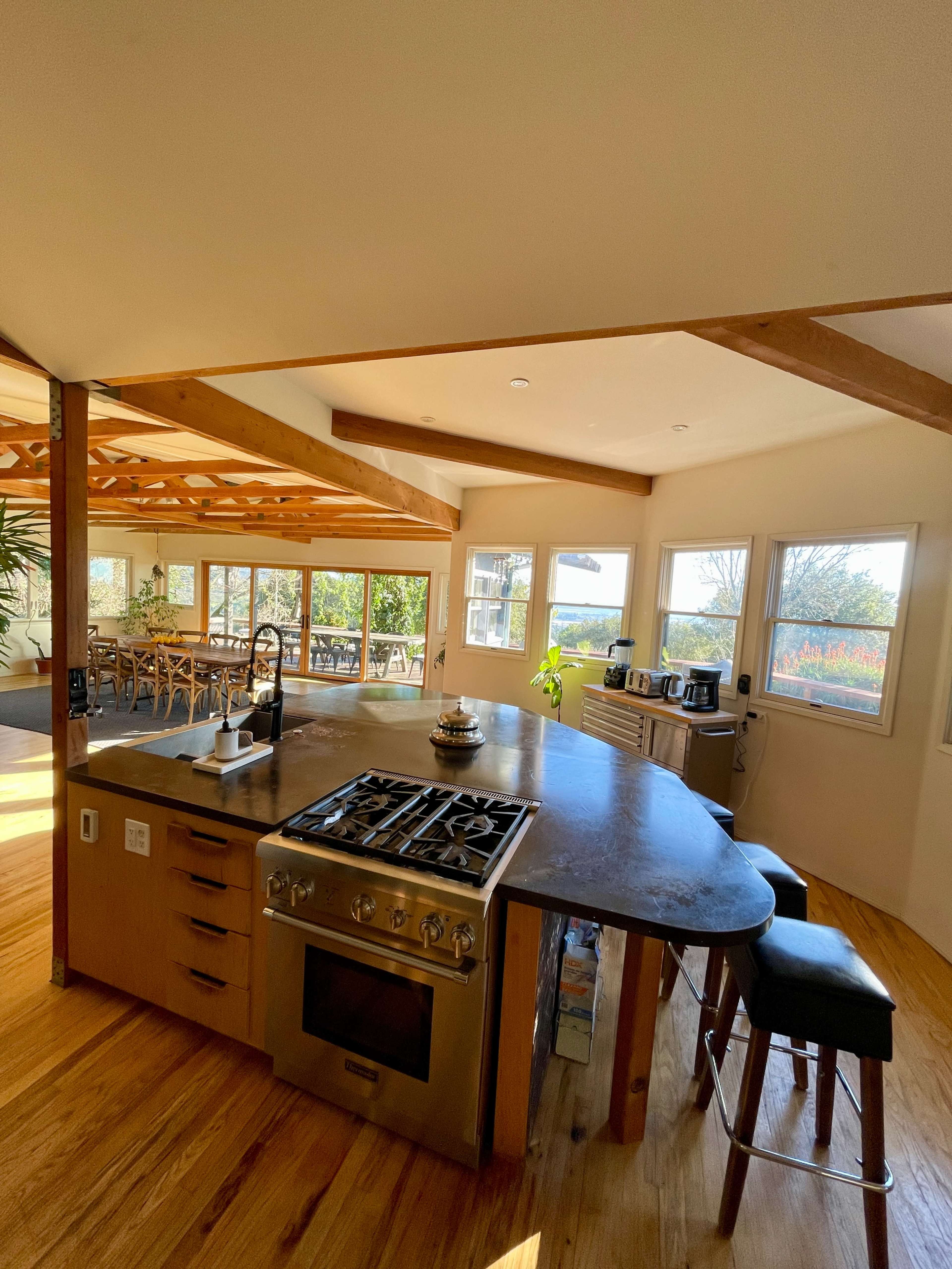 The image shows a modern kitchen with a curved countertop, stainless steel appliances, and large windows overlooking a scenic view.