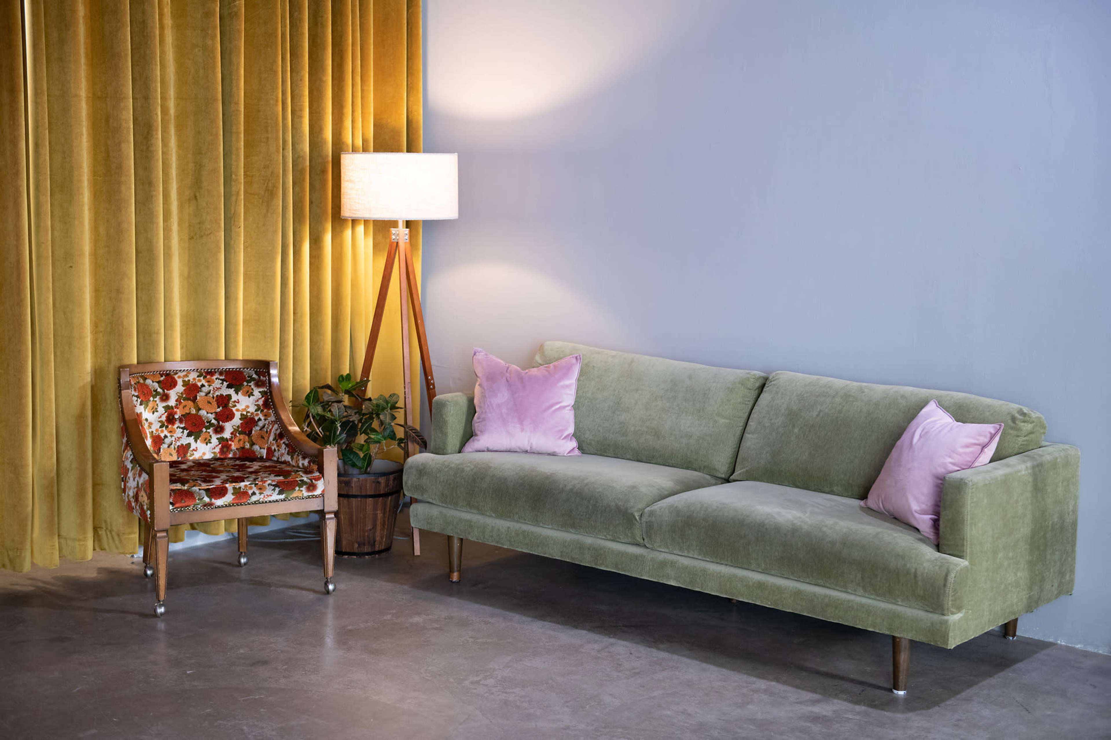 A green velvet sofa with pink cushions is positioned next to a patterned armchair and a tall lamp against a backdrop of yellow curtains and a grey wall.