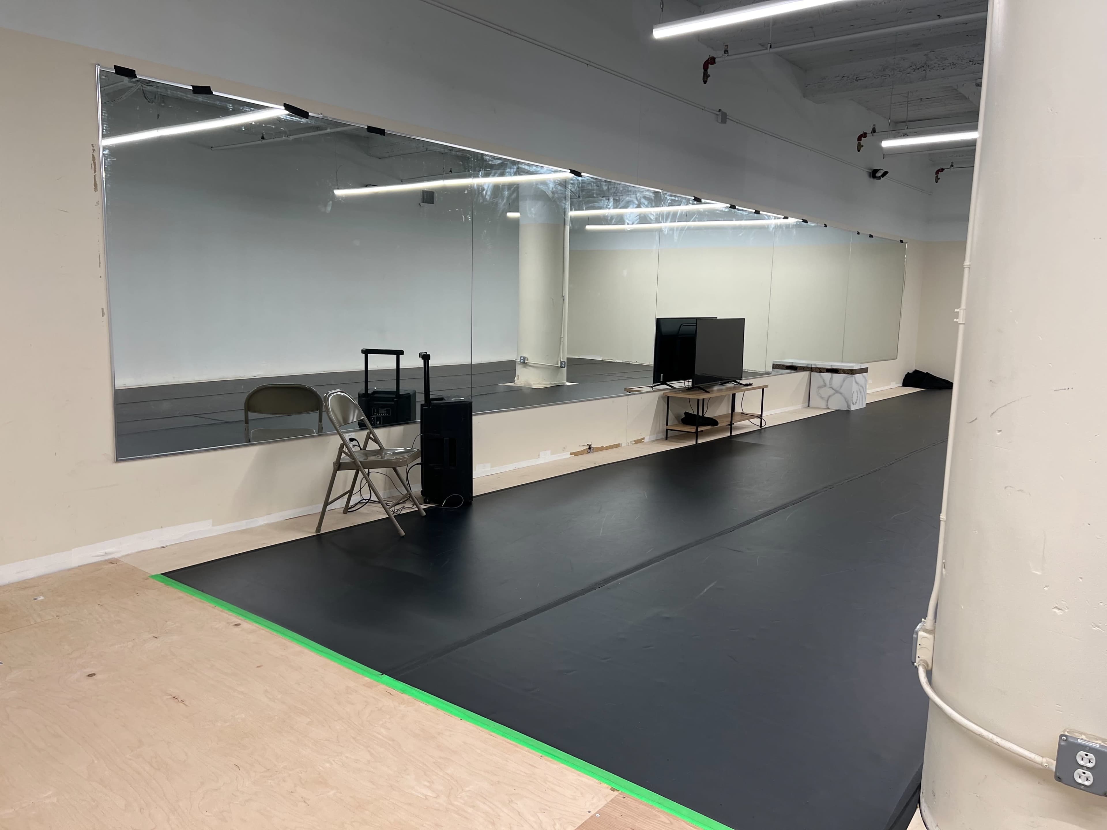 An empty dance studio with a large mirror, a television, a folding chair, and a black floor mat.
