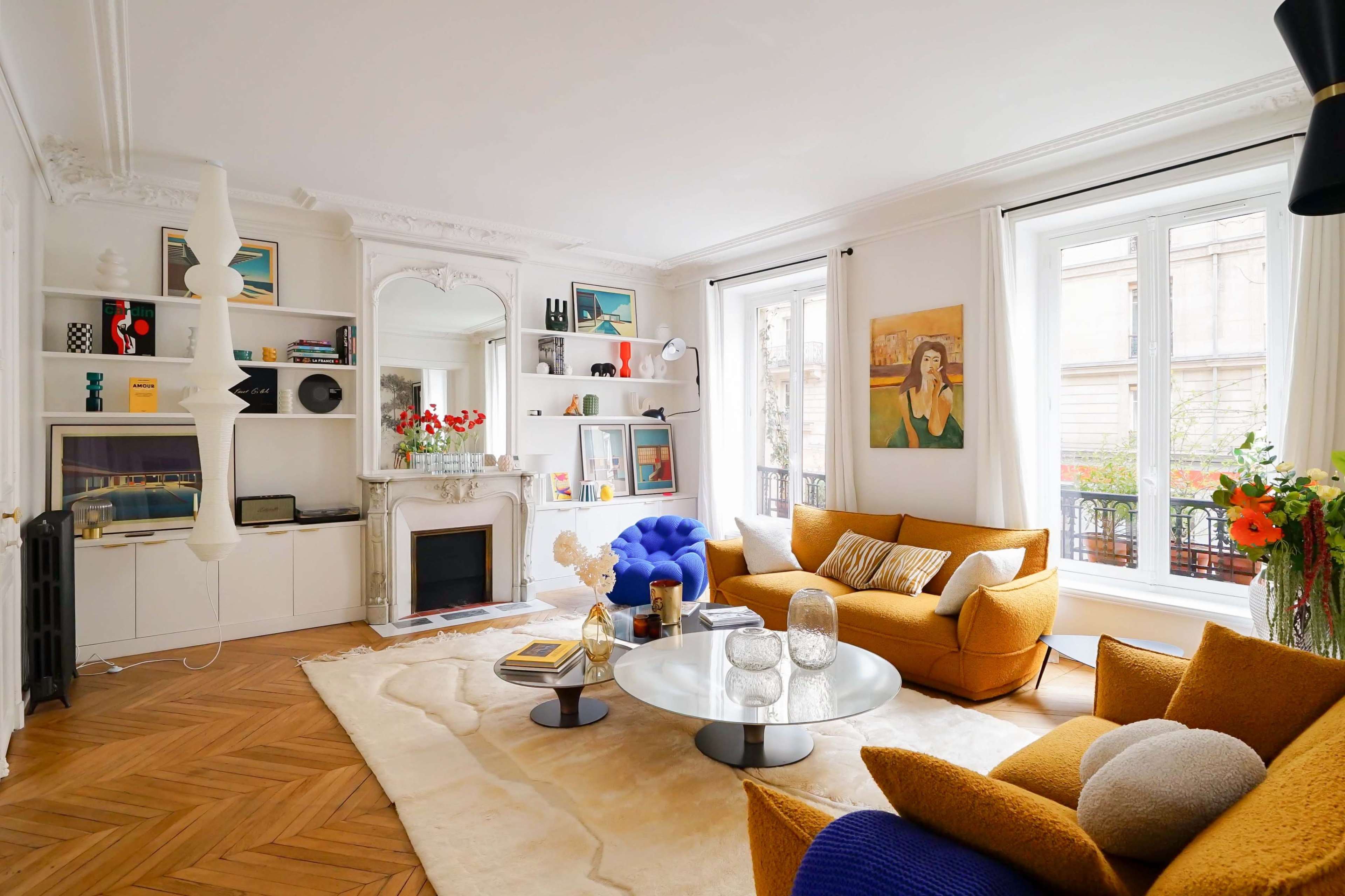 The living room features a combination of a yellow sofa and a blue chair, a round coffee table, a decorative rug, and large windows that provide natural light.