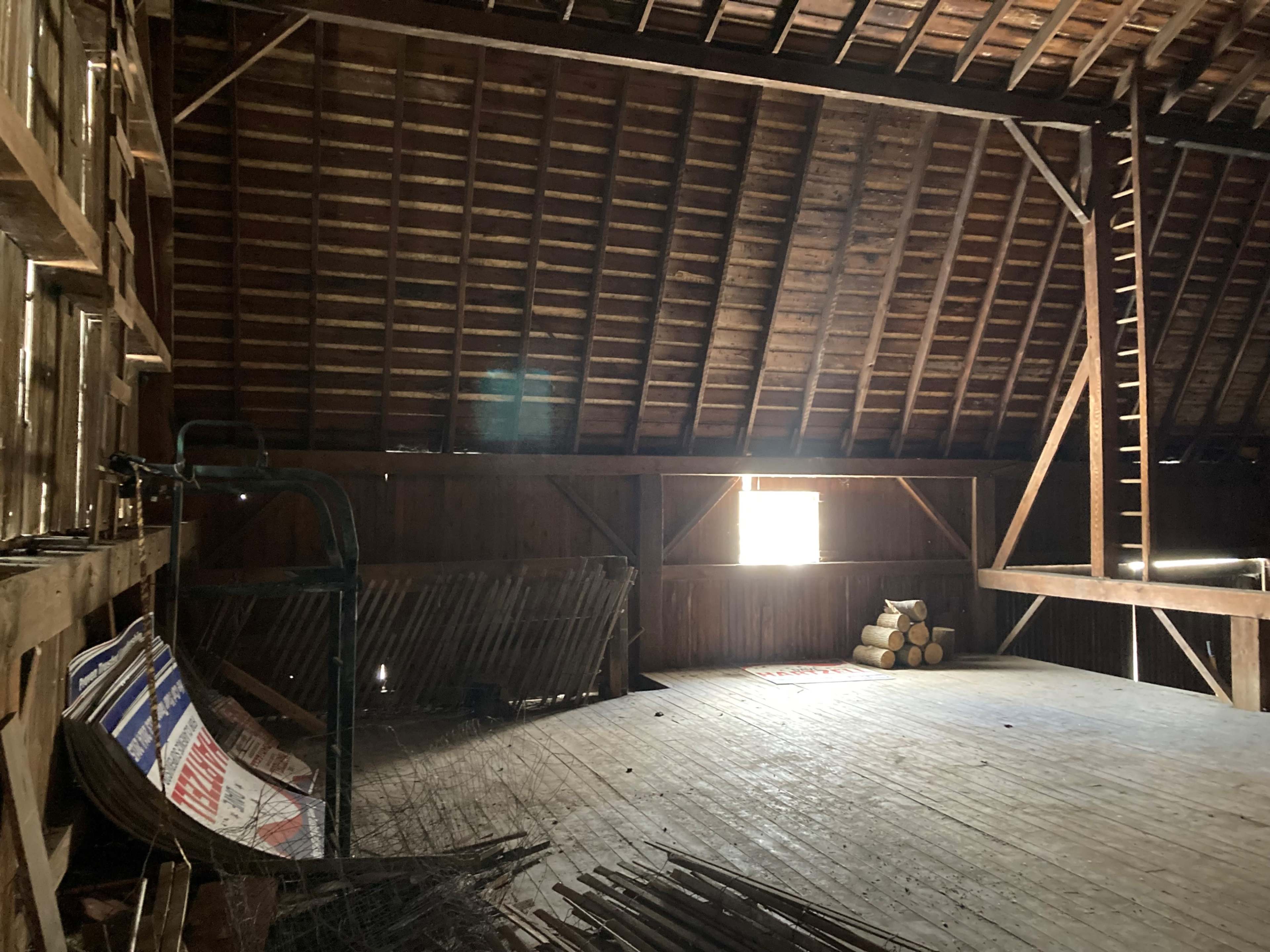 The interior of an empty barn with wooden beams, a loft area, and light coming through a window.