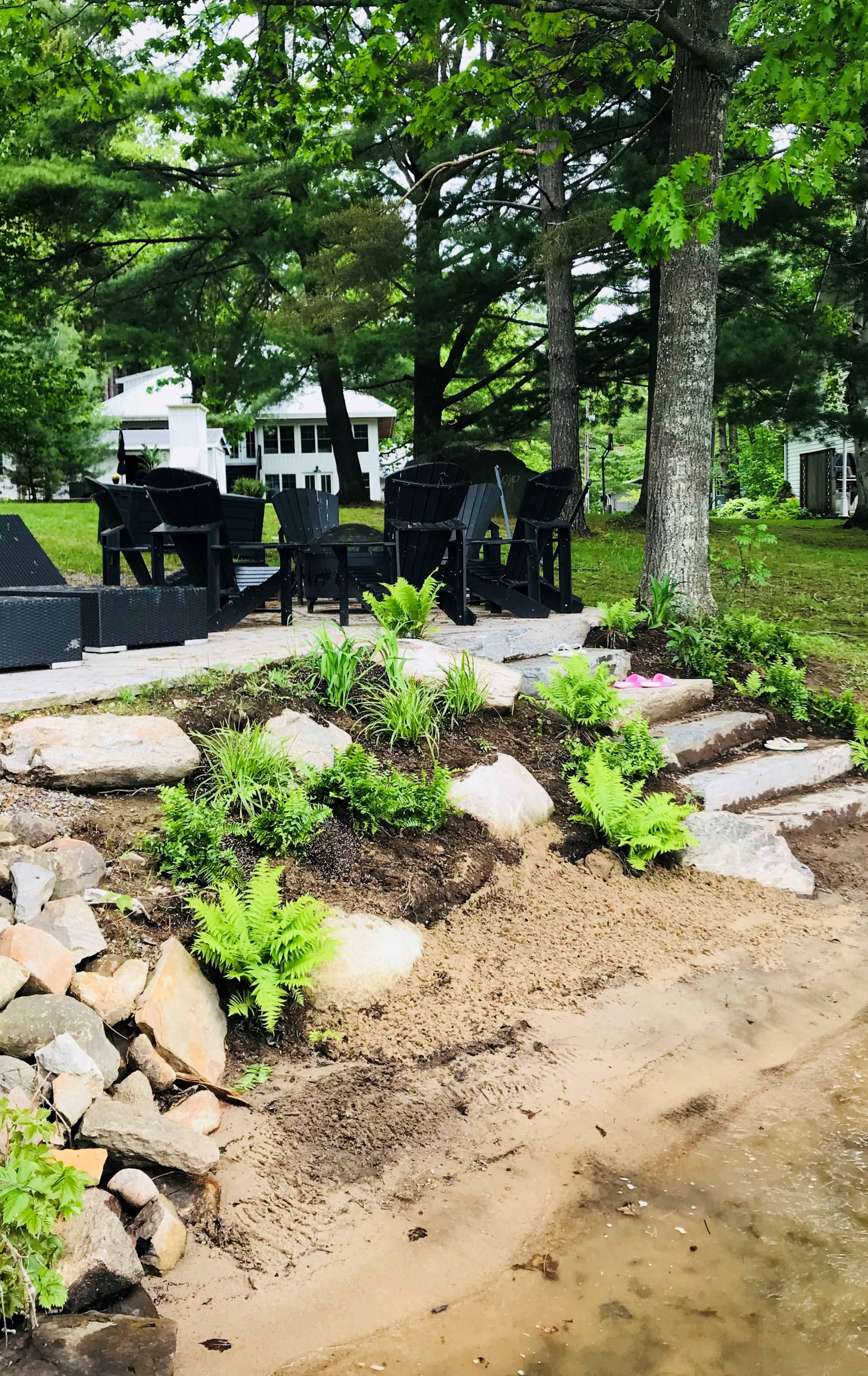 A landscaped area by a water's edge featuring stone steps leading down to a sandy spot, with black Adirondack chairs arranged on a grassy bank surrounded by ferns and stones.