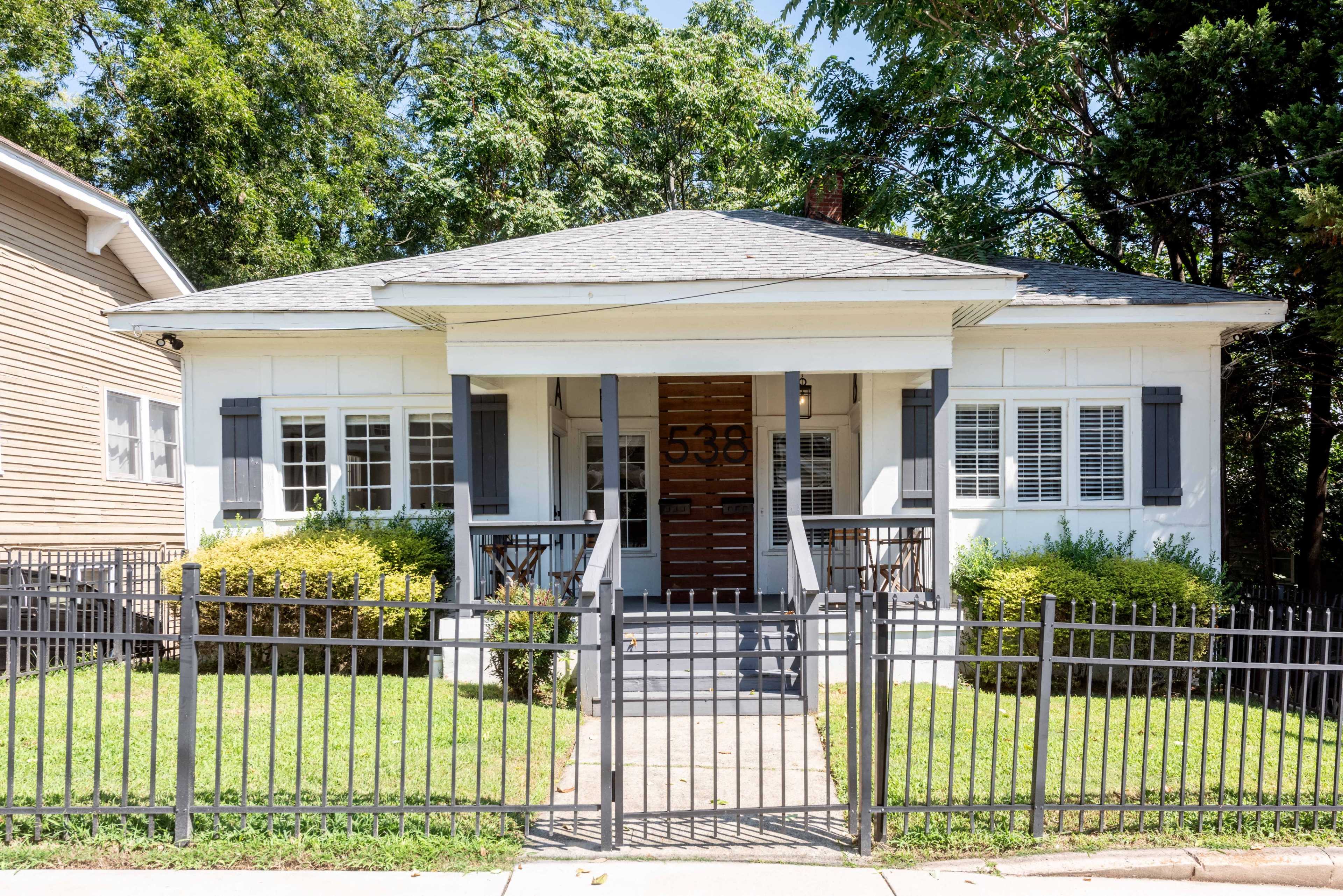 The image shows a single-story white house with a front porch, a brown door, and a black wrought iron fence surrounding the well-maintained lawn.