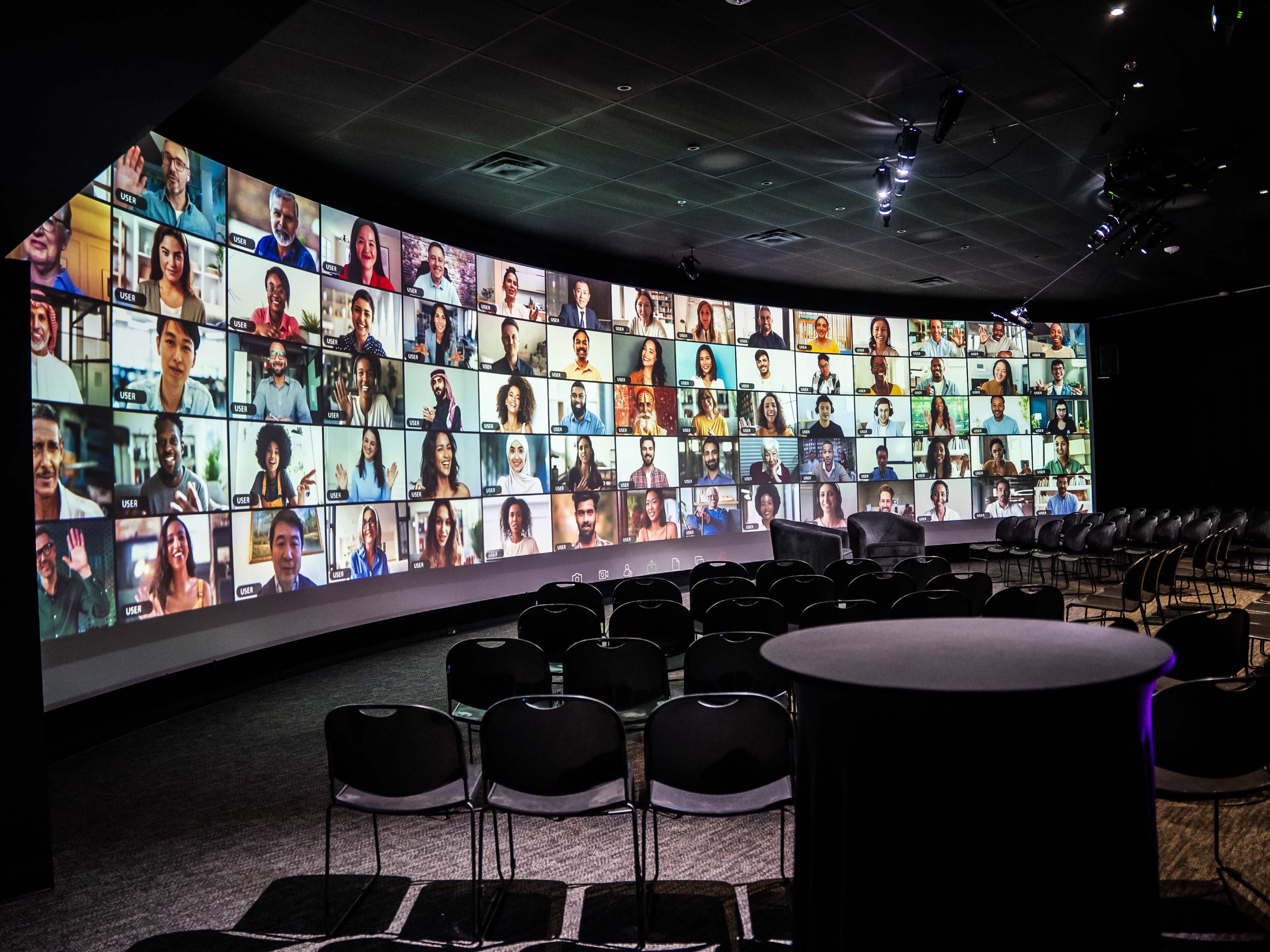 A curved screen displays a grid of diverse individuals participating in a virtual meeting, while empty chairs are arranged in the foreground.