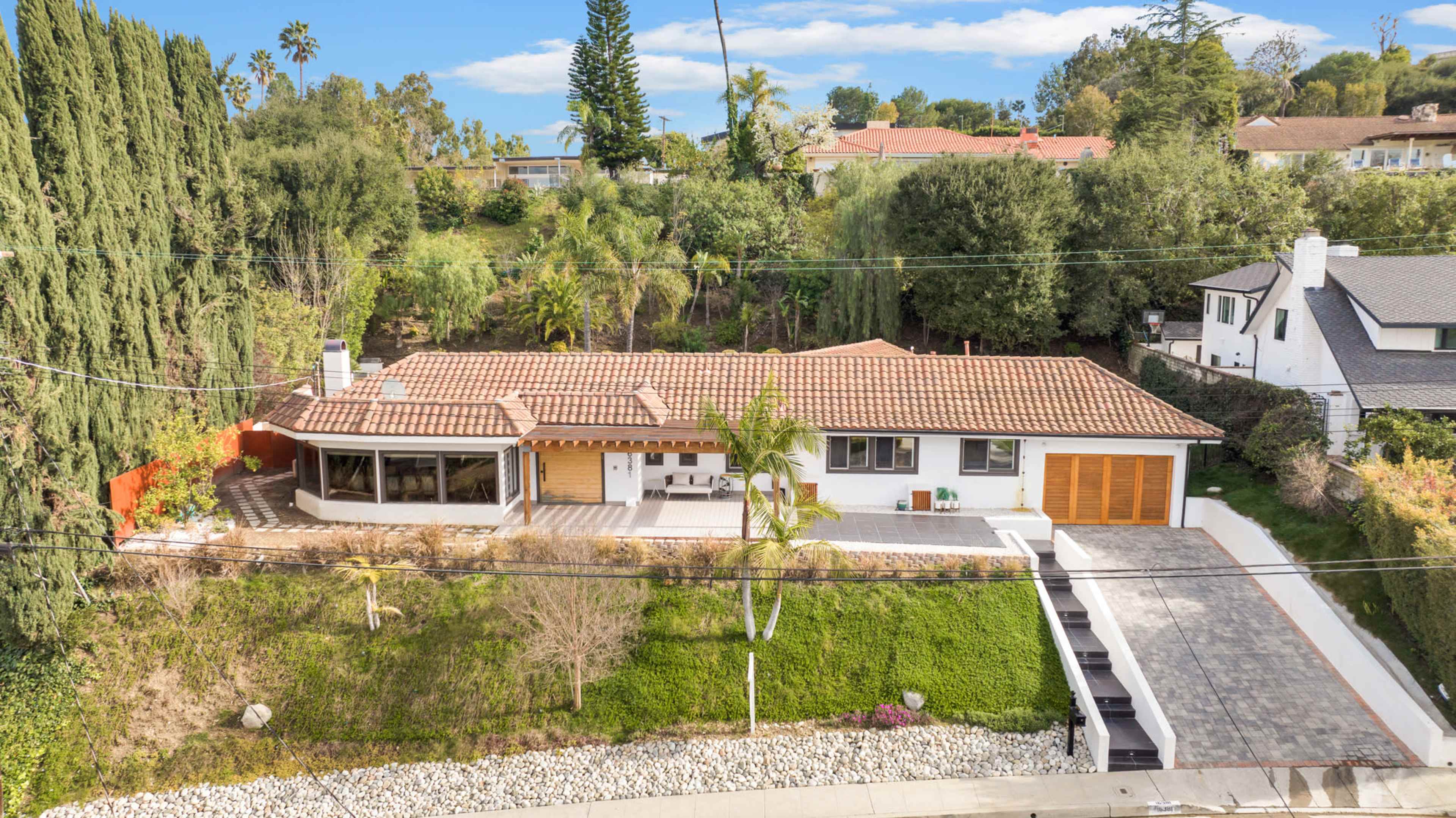 A single-story, modern home with a tiled roof sits on a landscaped hillside, featuring a driveway and a patio area surrounded by greenery.