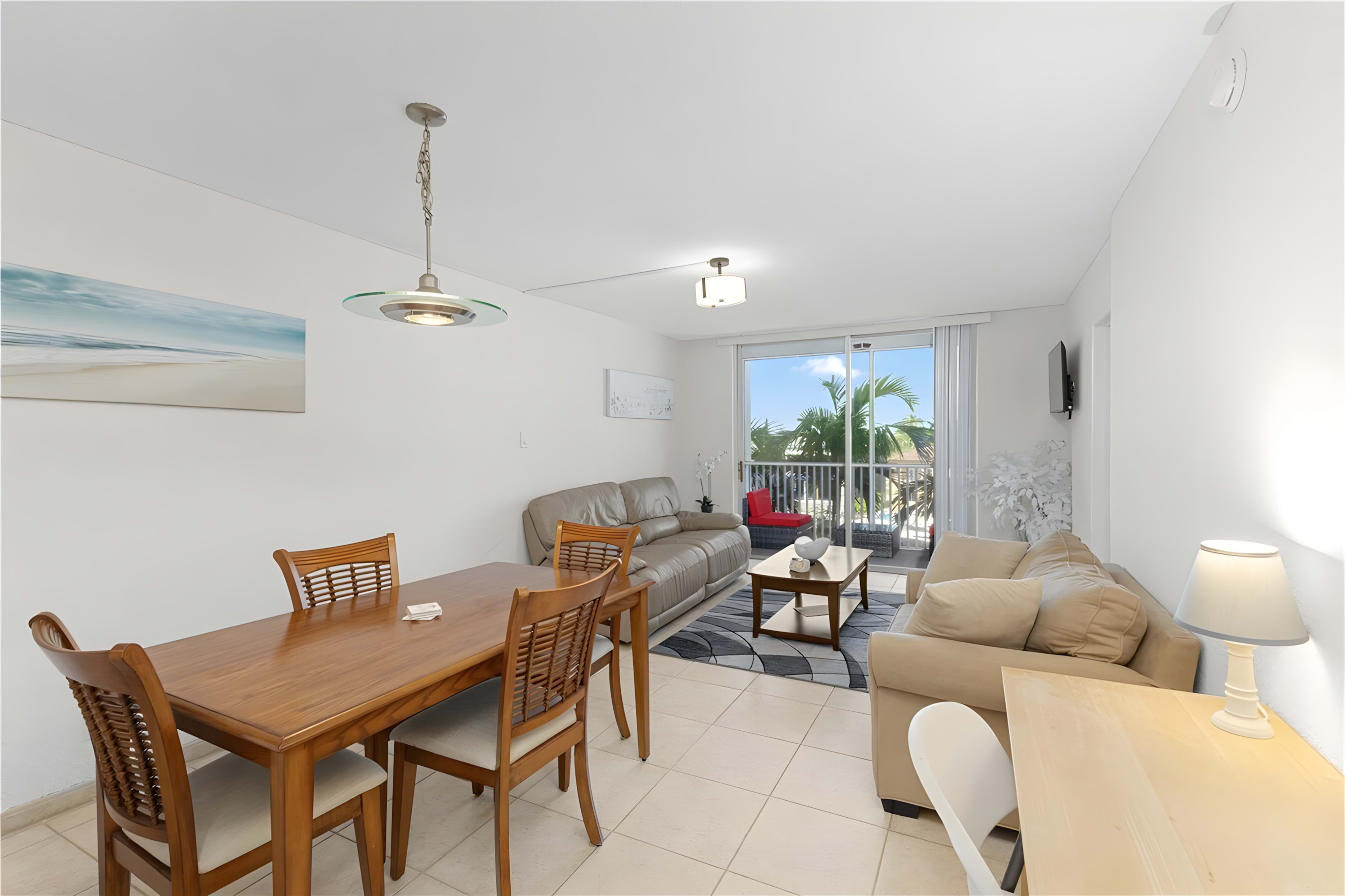 The image shows a modern living area with a dining table, seating arrangements, and a large window overlooking greenery.
