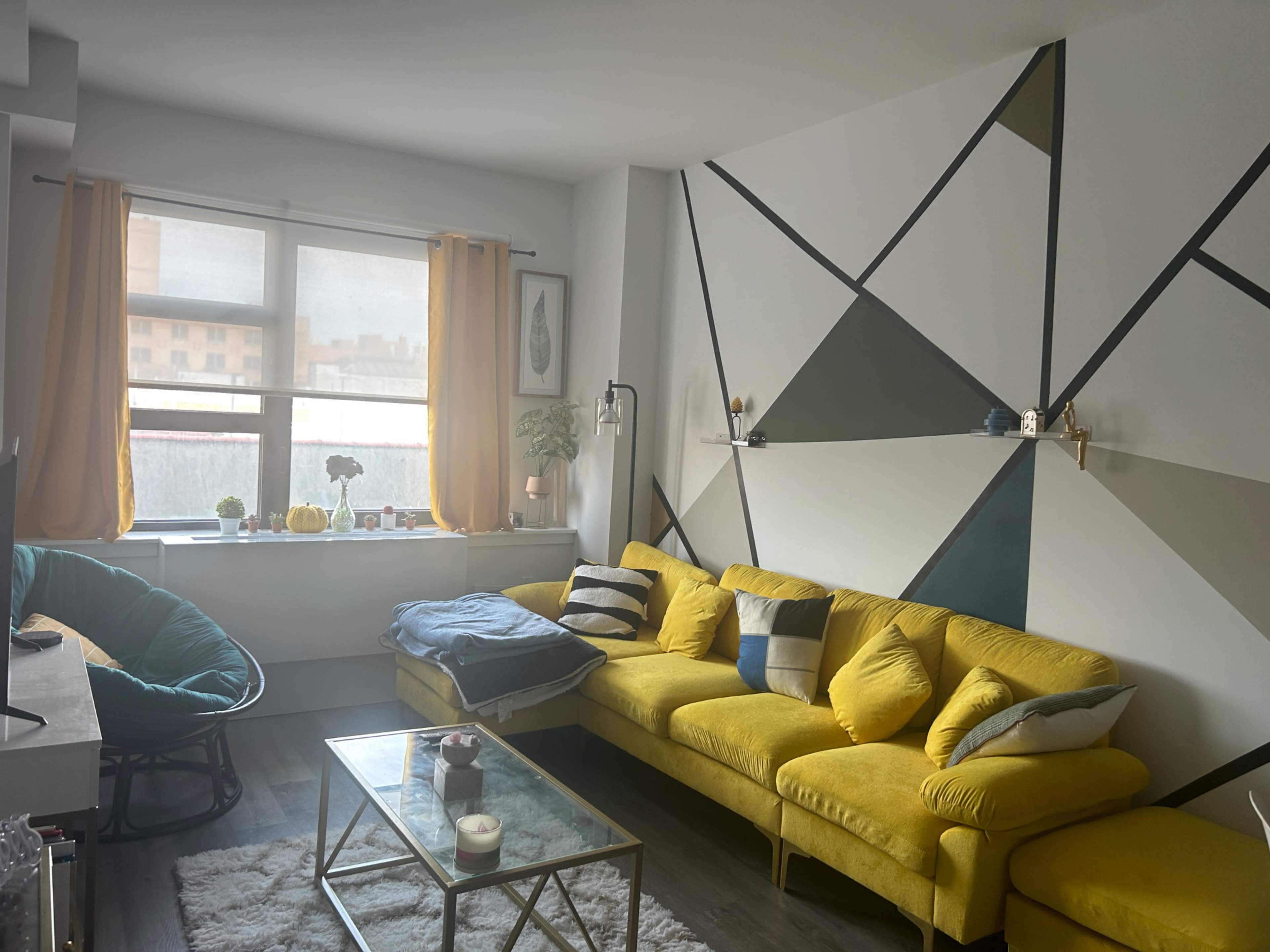 The image depicts a modern living room with a yellow sectional sofa, a glass coffee table, a large window, and geometric wall art.