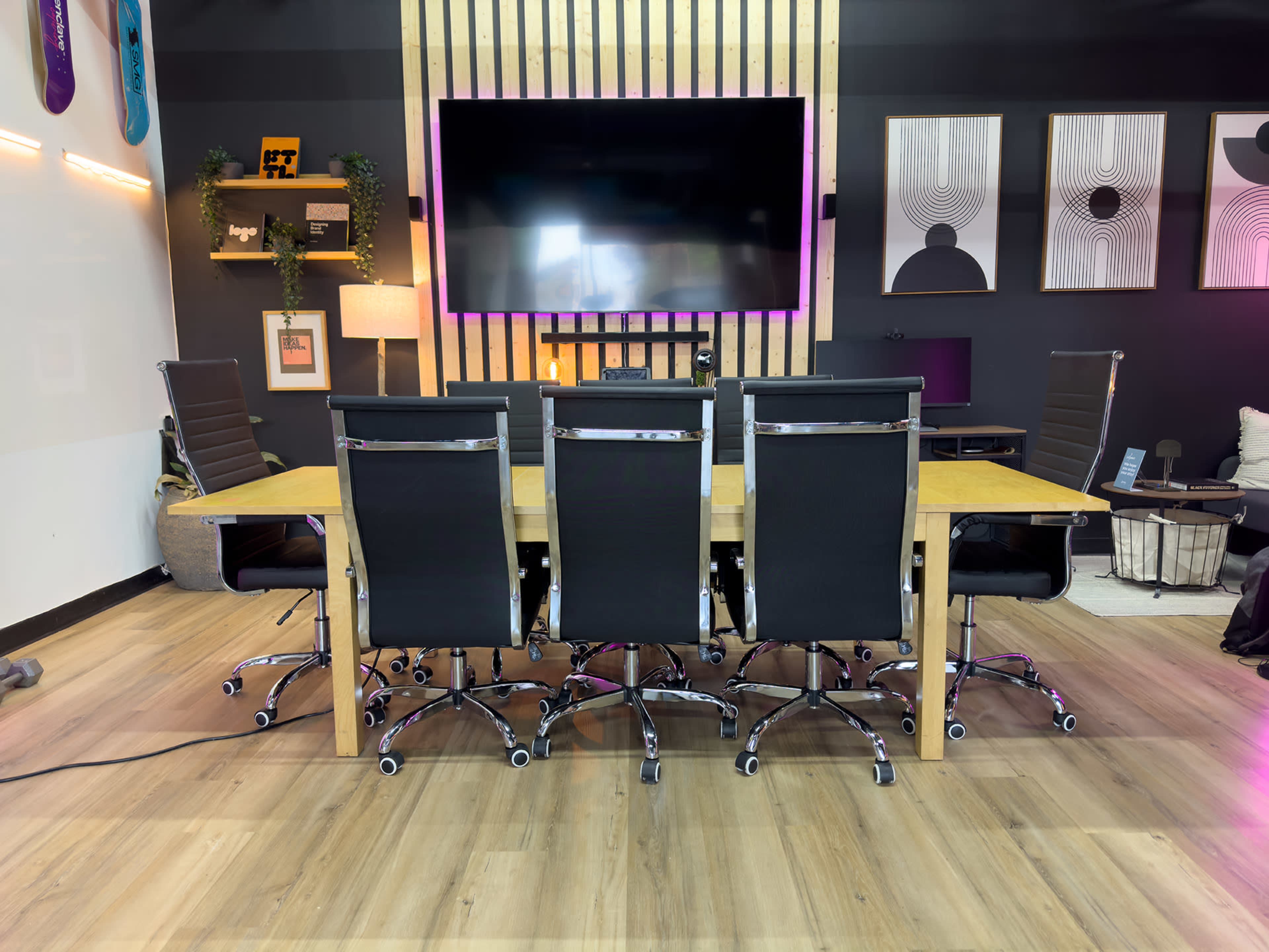 A modern conference room features a long wooden table surrounded by ergonomic black chairs, with a large screen mounted on the wall and various decorative elements.