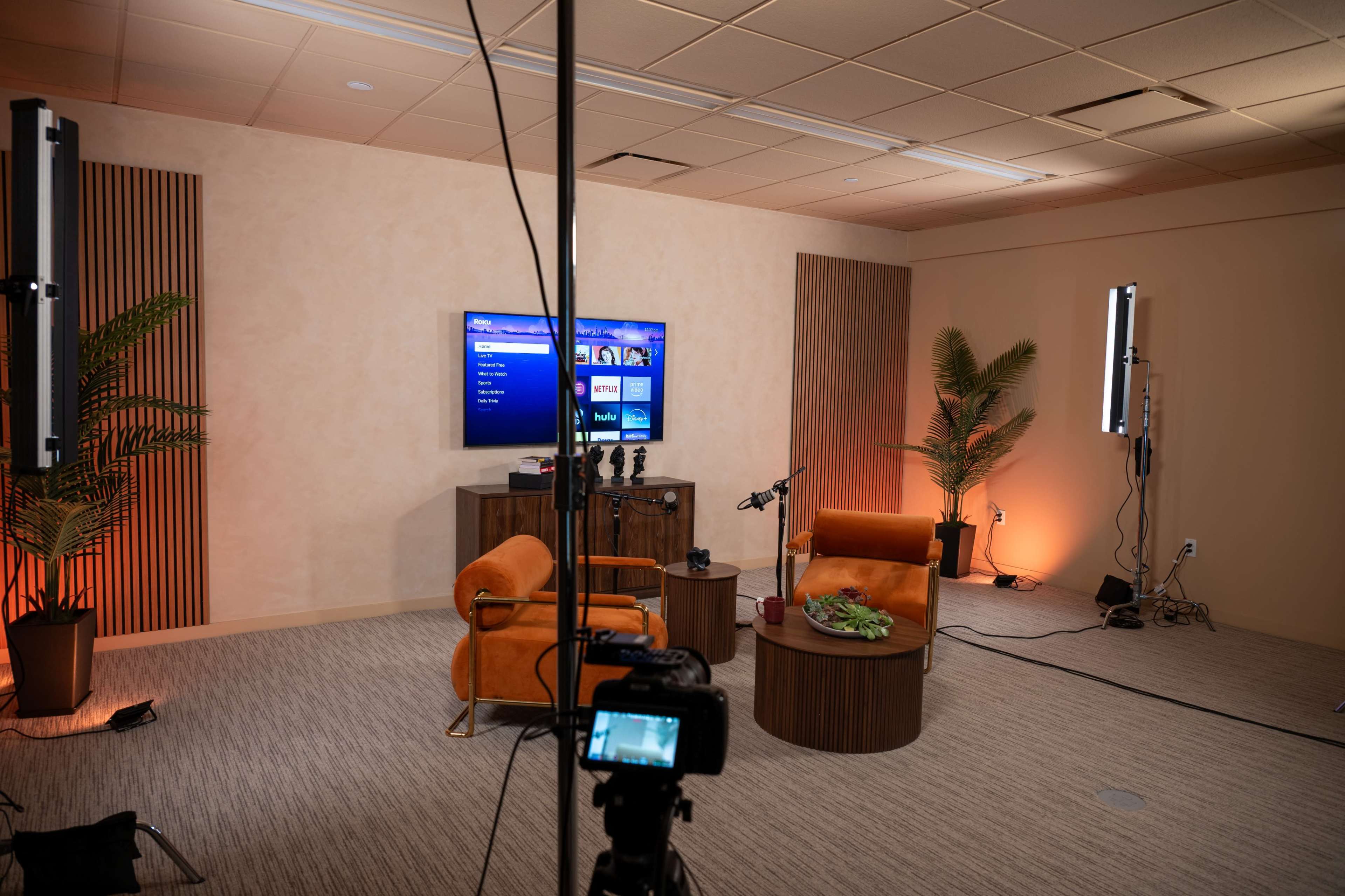 A media studio setup features two orange chairs facing a television screen, with plants and recording equipment arranged around the space.