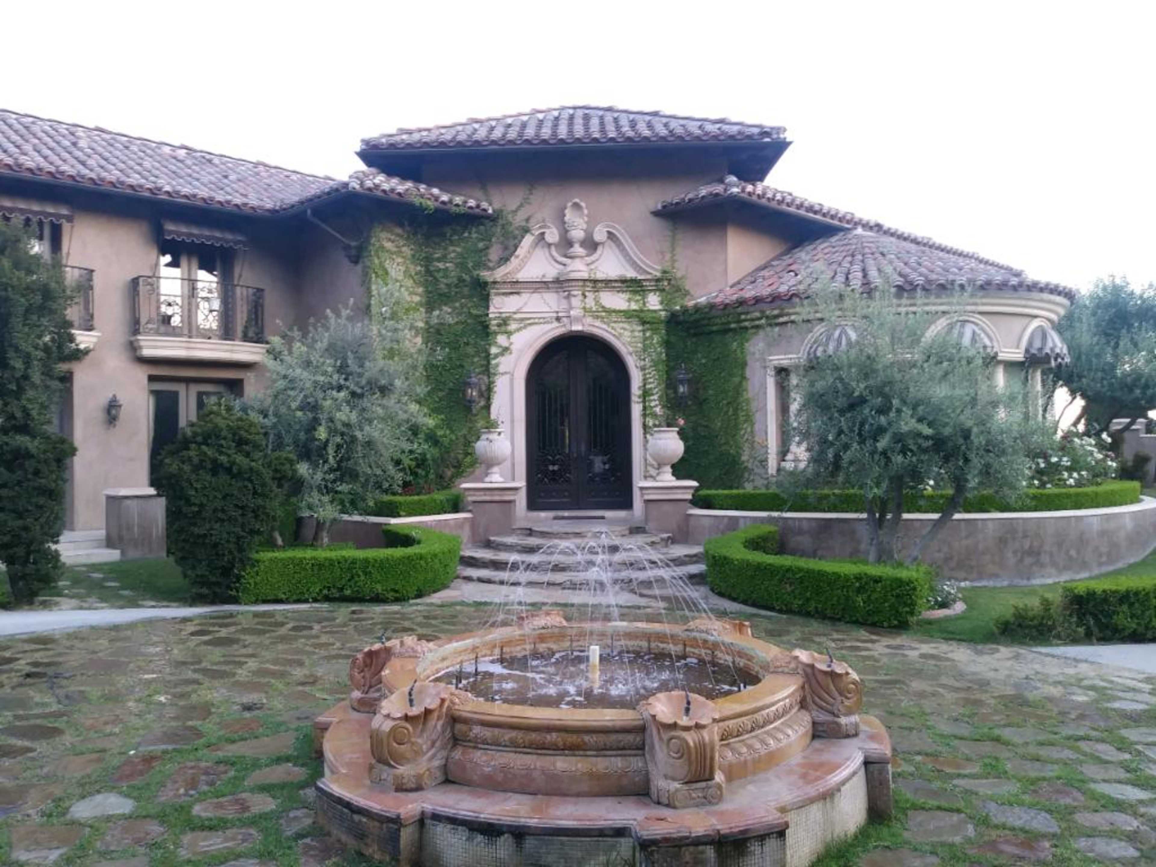 The image shows a luxurious home with a grand entrance, surrounded by manicured landscaping and a central fountain.