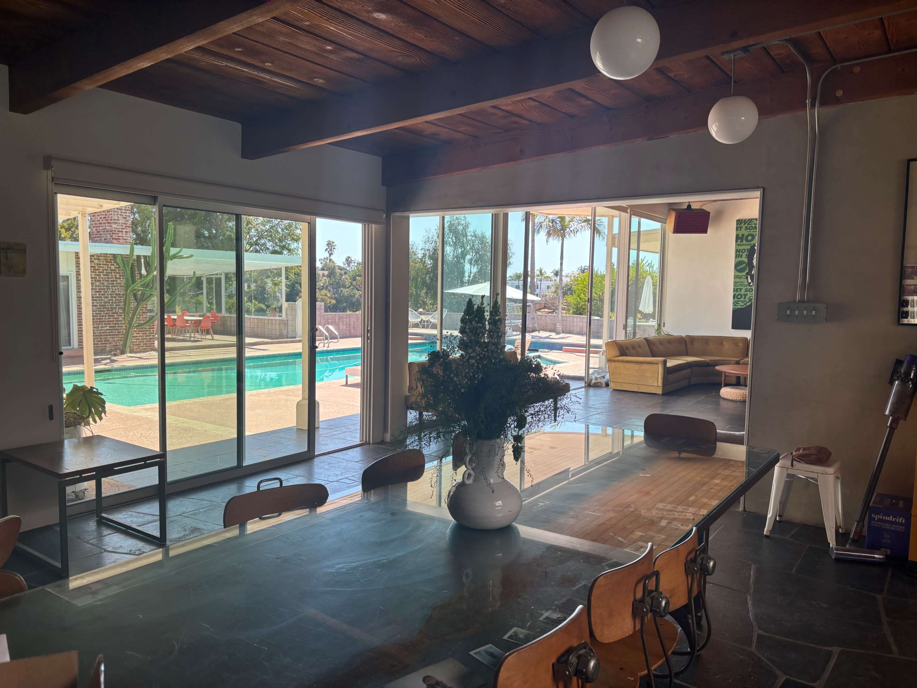 The image shows a modern interior space with a dining table, a view of a swimming pool through large glass doors, and a cozy seating area visible in the background.