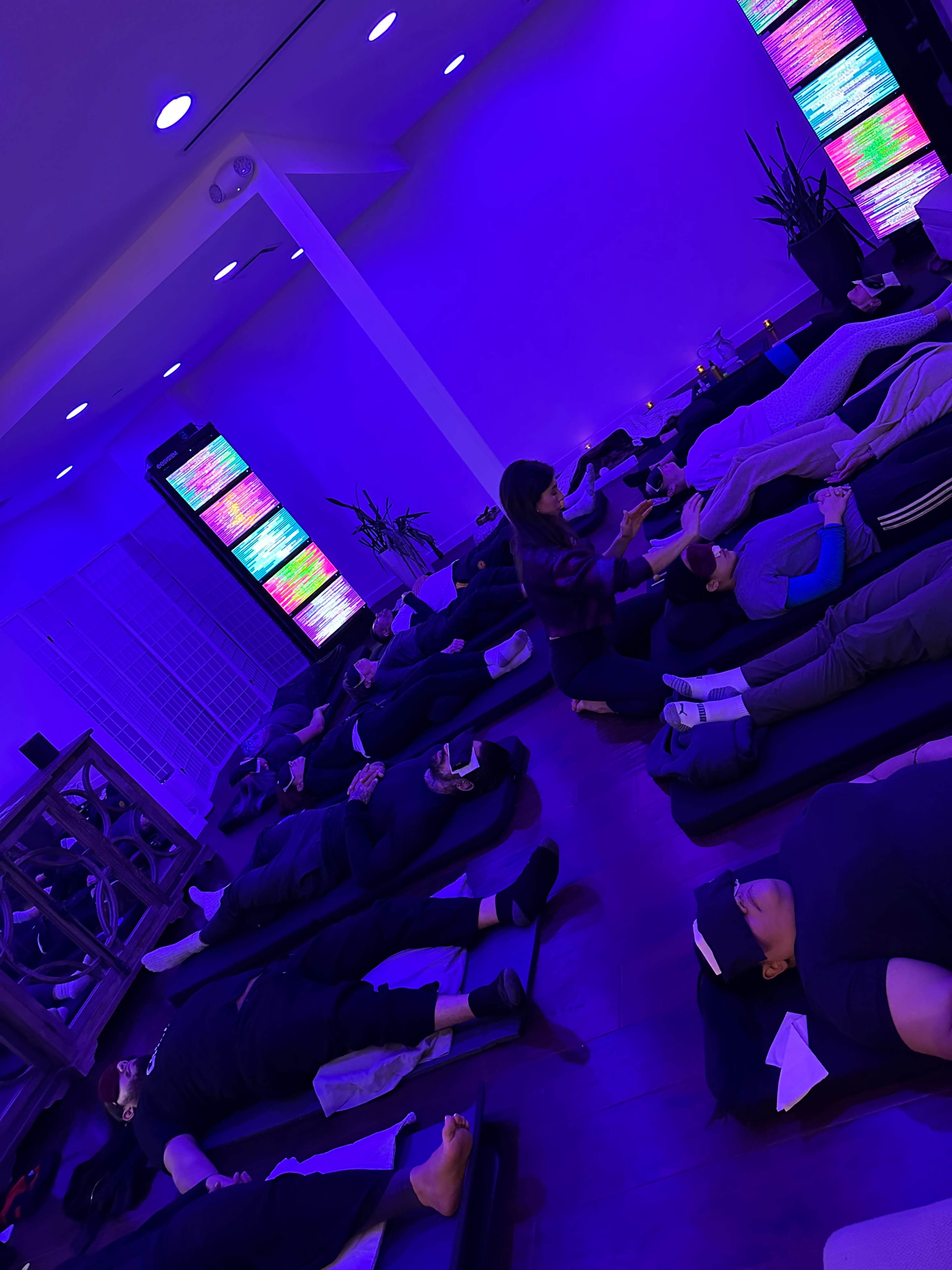 A group of people lie on mats in a dimly lit room with purple lighting and large screens displaying colorful patterns.