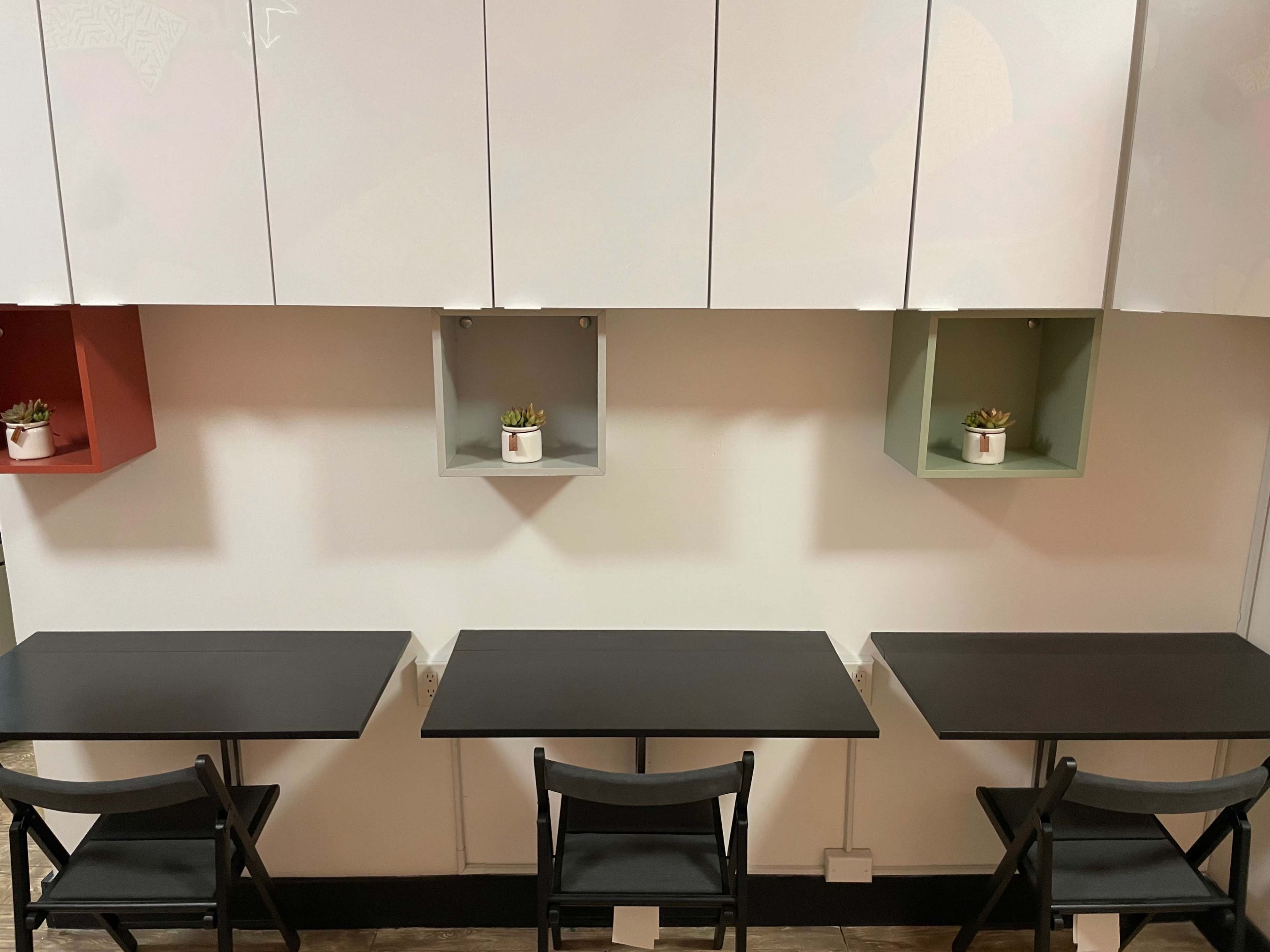 The image shows three black tables with chairs arranged against a wall, featuring three small plants in colorful wall-mounted shelves above them.