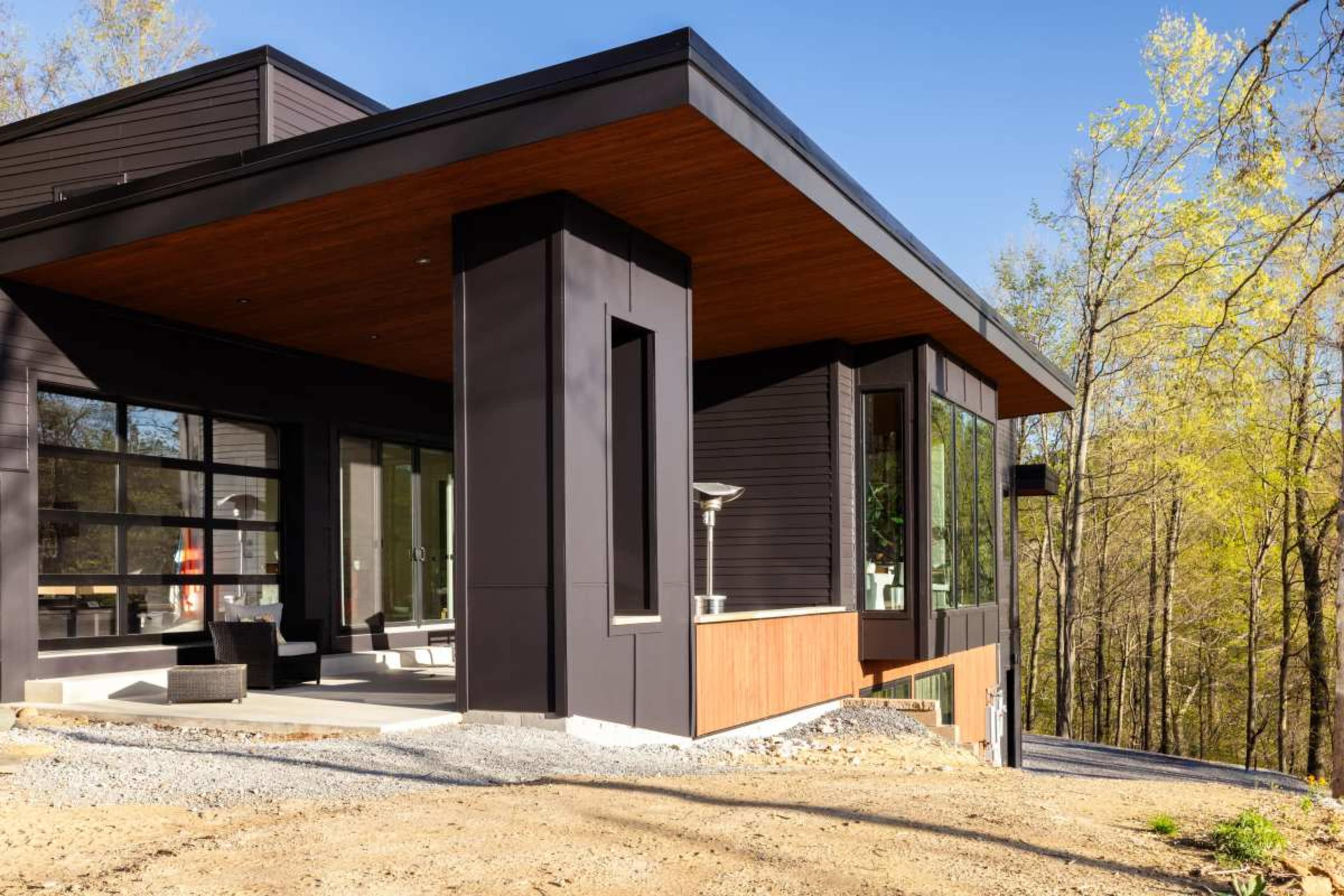 A modern house featuring a combination of dark exterior materials and large windows, situated near a wooded area with sparse foliage.