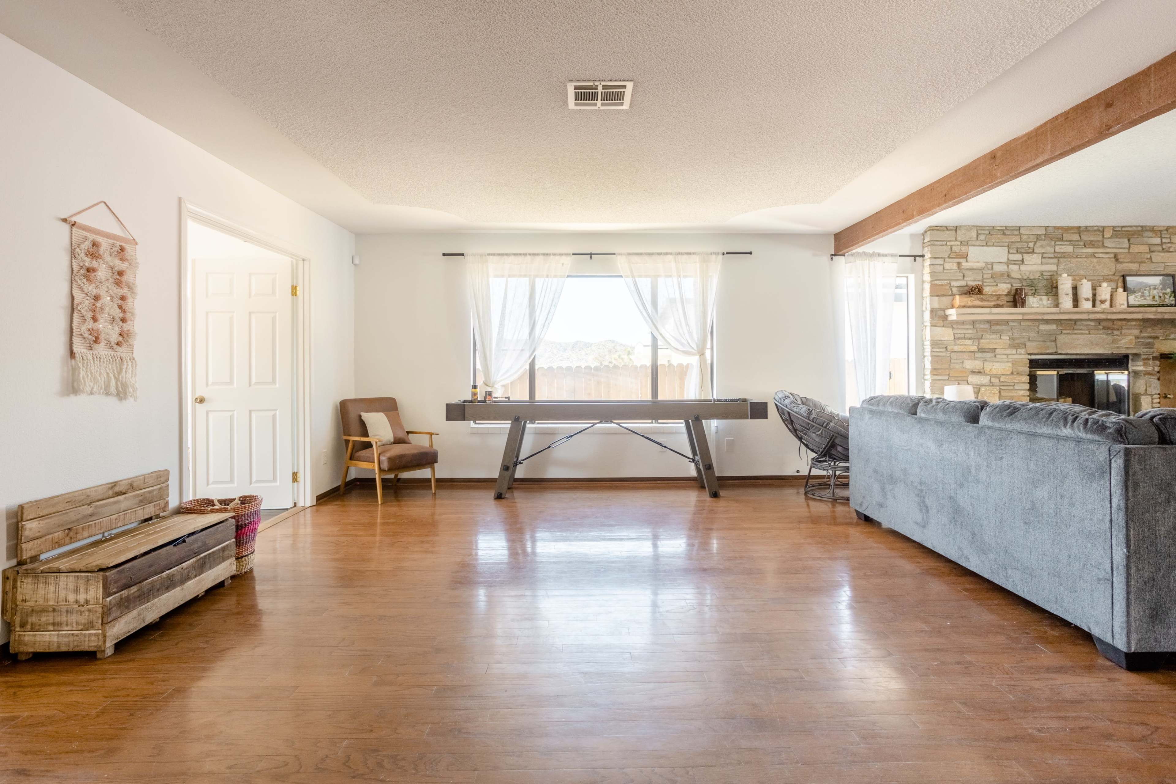 A spacious living room with wooden floors, a large window with sheer curtains, and a stone fireplace, featuring a ping pong table in the center of the room.