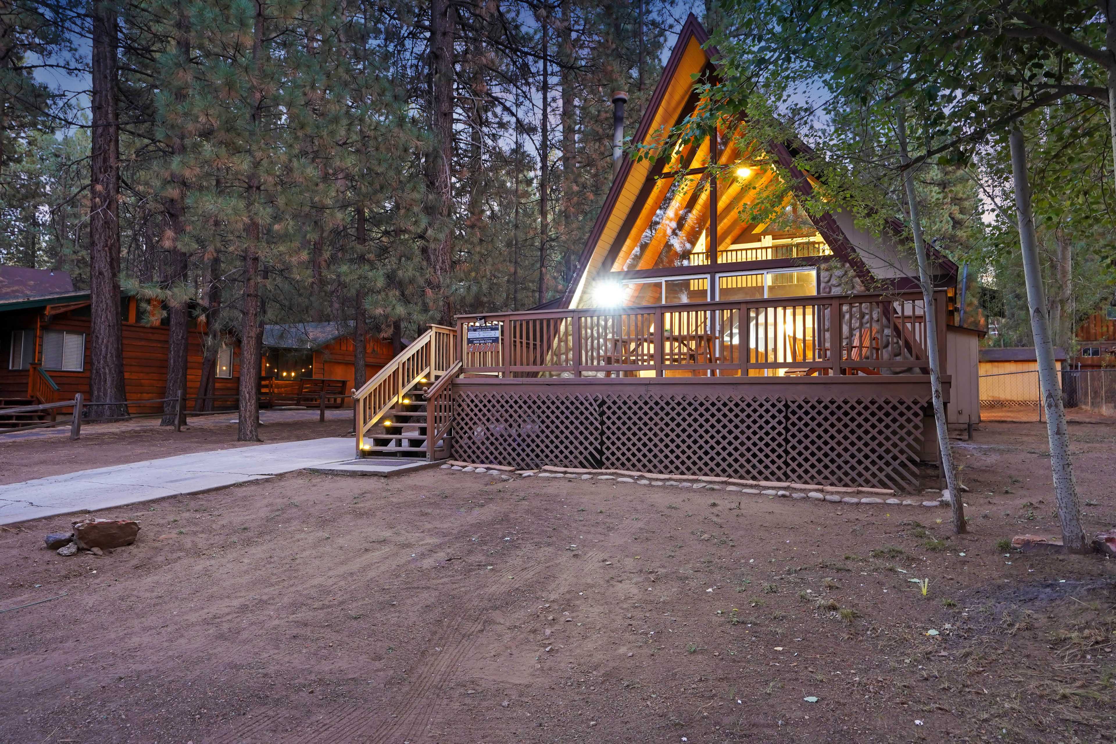 A wooden A-frame cabin is illuminated at dusk, surrounded by tall pine trees and gravel paths.