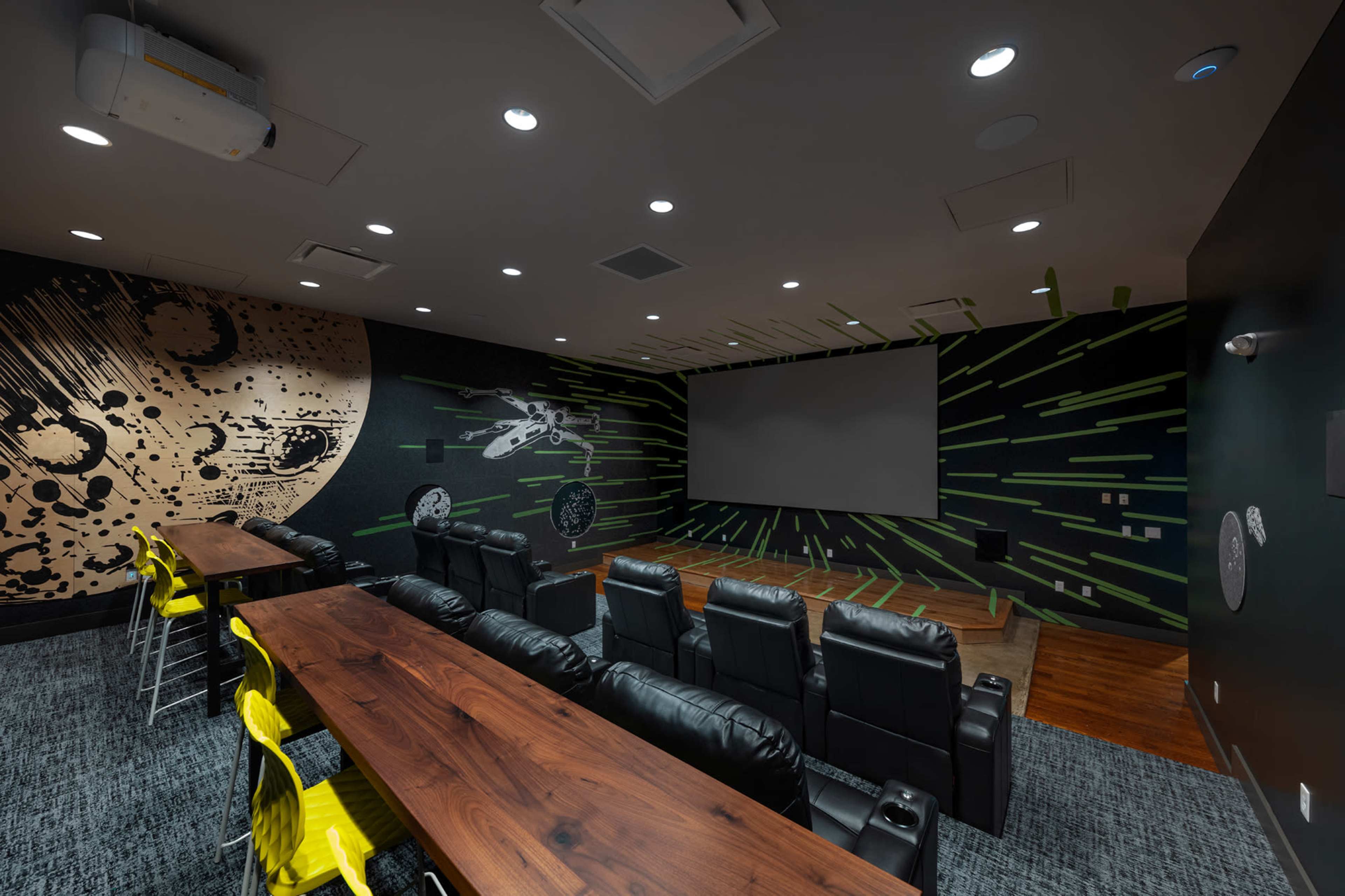 The image shows a modern home theater with a large projection screen, leather recliner seats, and a decorative wall featuring a space-themed mural.