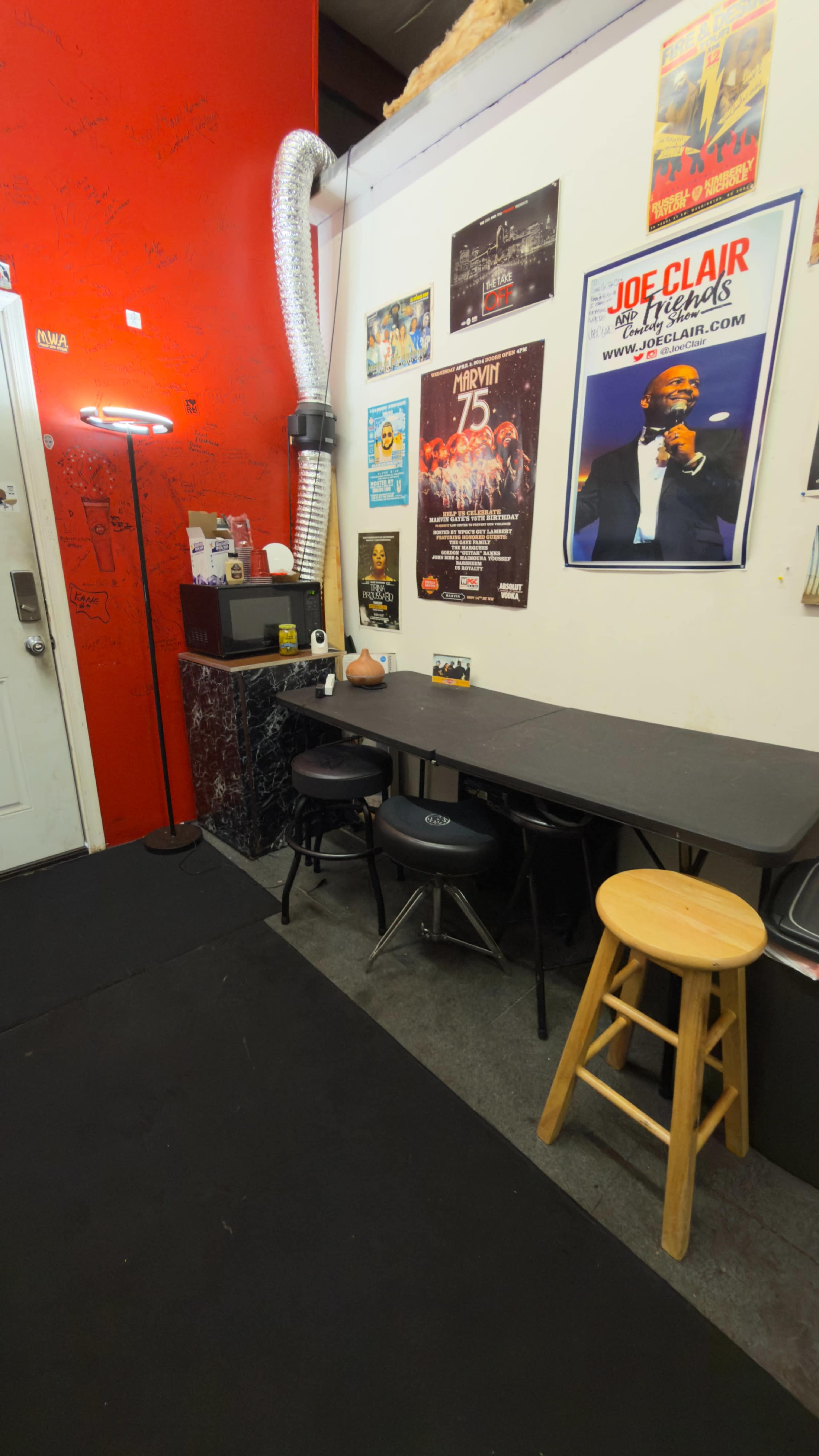 The image shows a small room with a red wall, a black table, several chairs, and various posters on the wall.