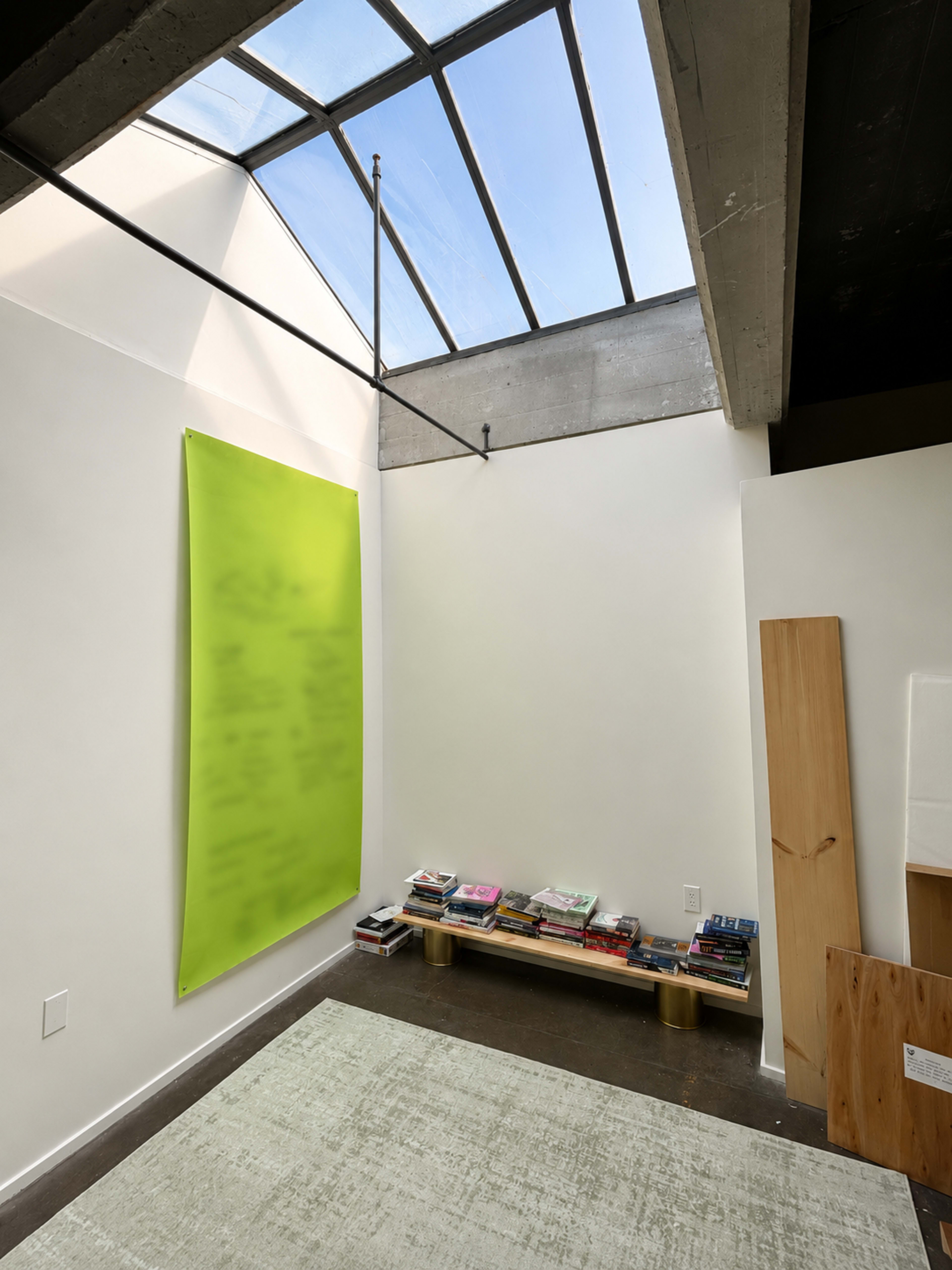 The image shows a minimalist interior space with a skylight, a green wall poster, a low wooden shelf stacked with books, and a light green rug on the floor.