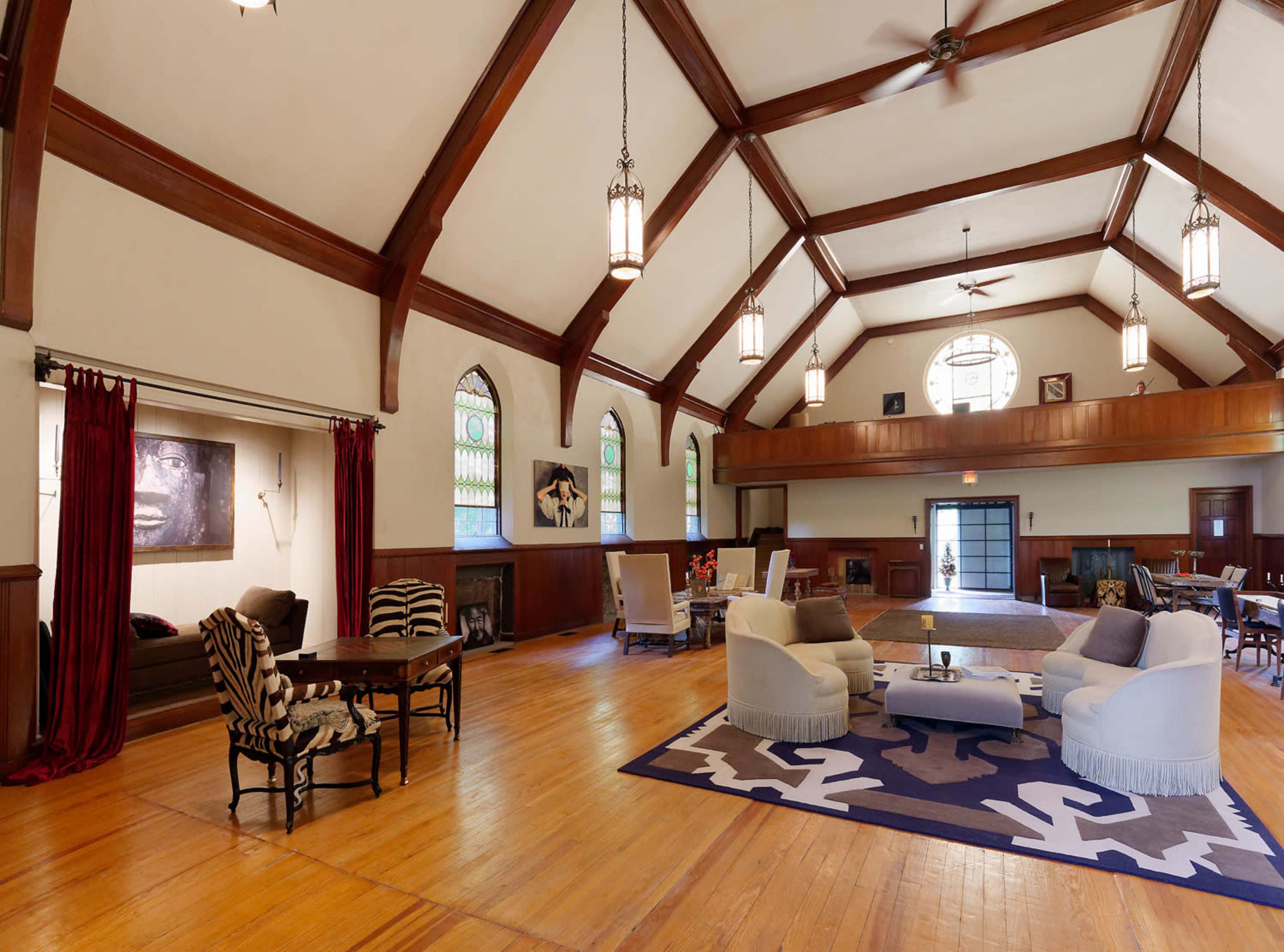 A spacious interior of a room with high wooden beams, large windows, and various seating arrangements, including a table and chairs.