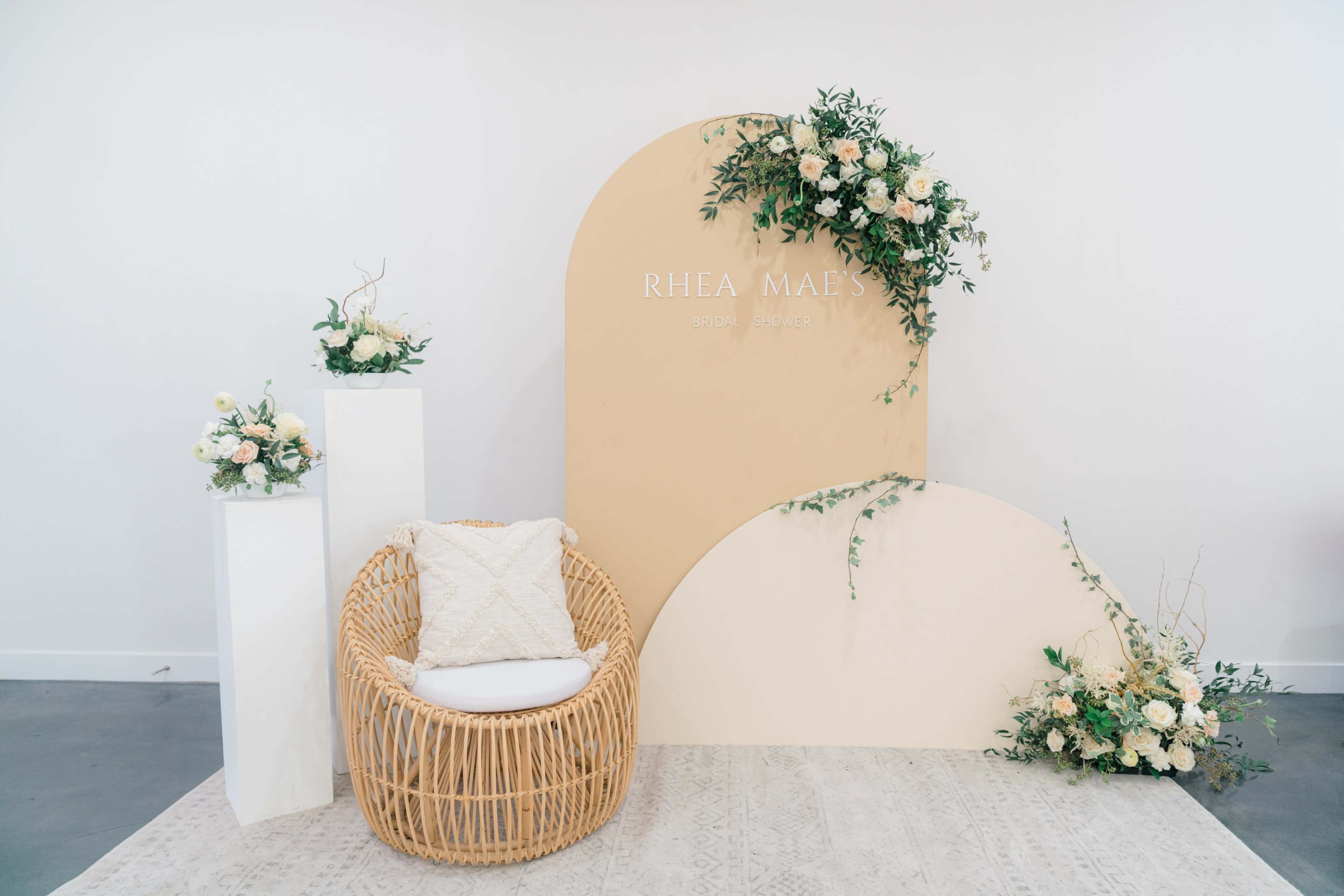 The image shows a simple event setup featuring a rattan chair, a backdrop with the name "Rhea Maes" in a minimalistic design, and floral arrangements on pedestals and the backdrop.
