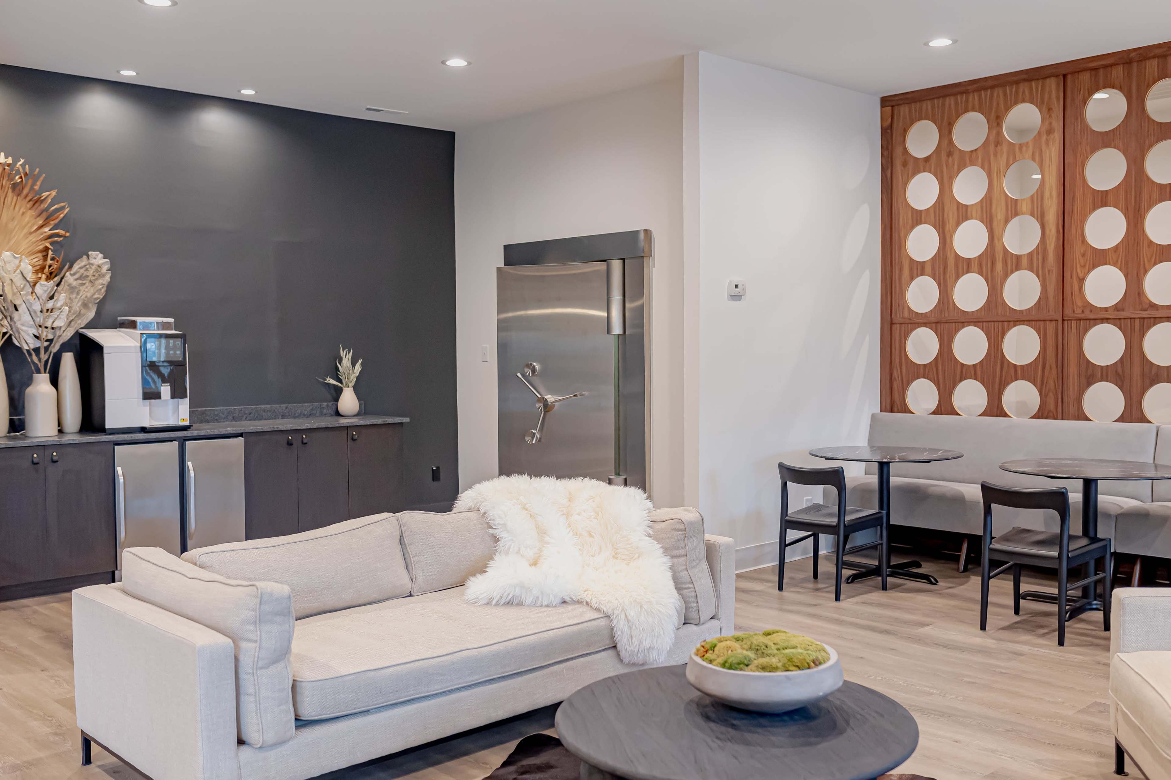 A modern living space featuring a light-colored sofa, a round black coffee table, a kitchen area with stainless steel appliances, and a dining area with round tables and minimalist seating.
