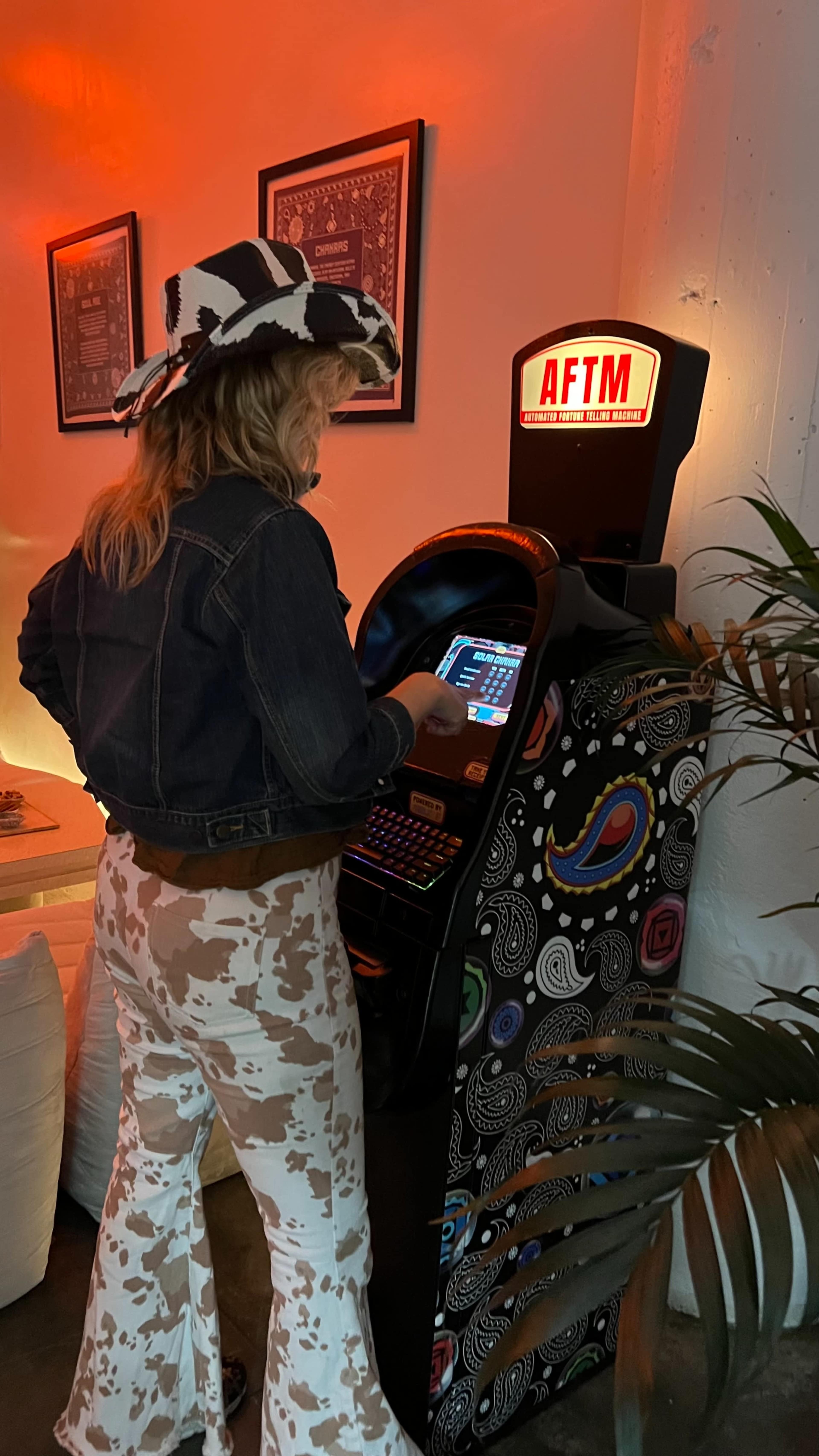 A person wearing a cowboy hat and denim jacket is standing in front of a colorful arcade machine, interacting with its screen in a dimly lit room.